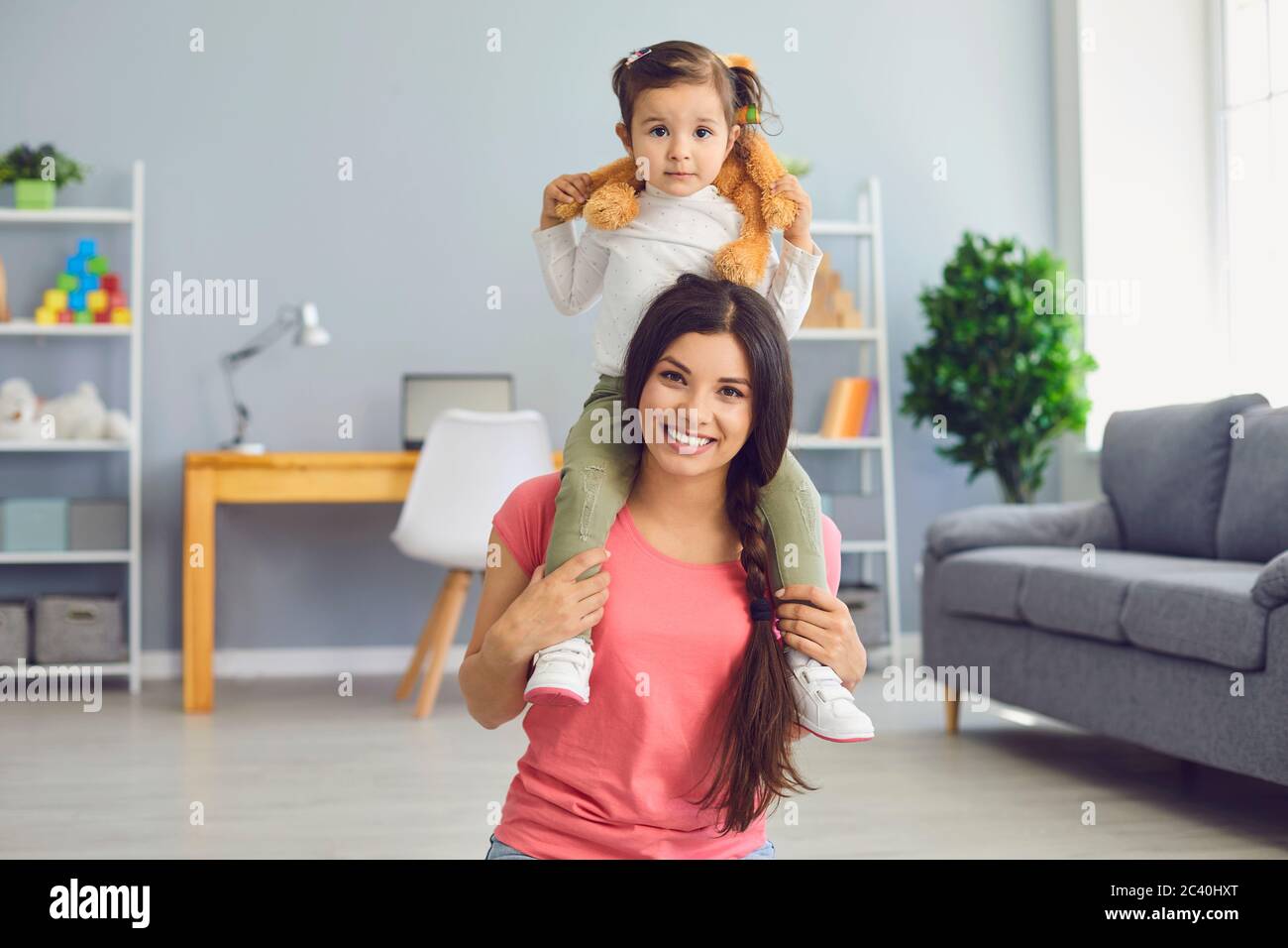 Une mère souriante portant une petite fille adorable sur ses épaules à la maison. Parent et enfant avec jouet jouant à l'intérieur Banque D'Images