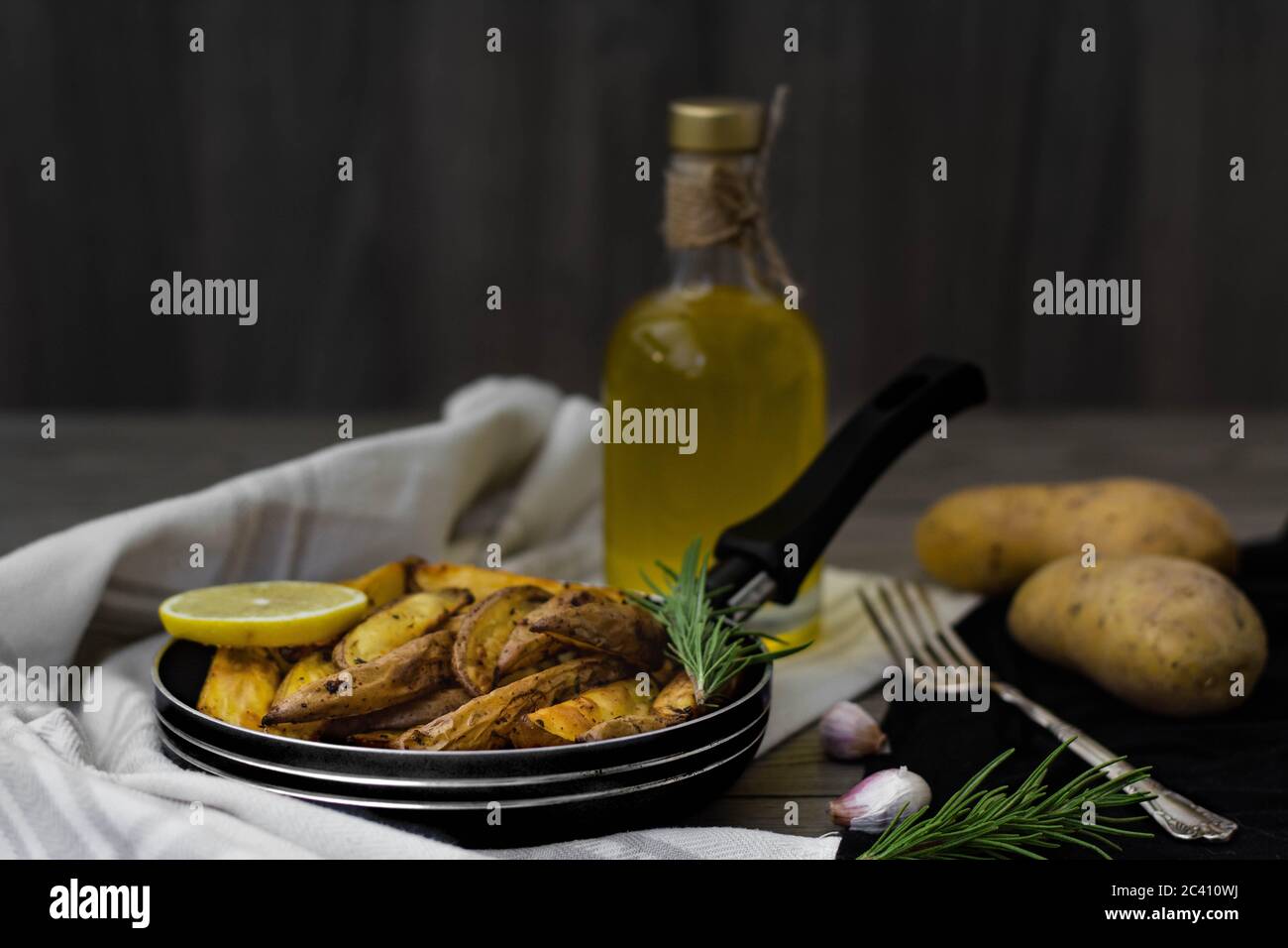 Quartiers de pommes de terre cuits au romarin, à l'ail et au citron dans une petite casserole. Pommes de terre crues, fourchette d'argent vintage, romarin et huile d'olive en arrière-plan. Banque D'Images