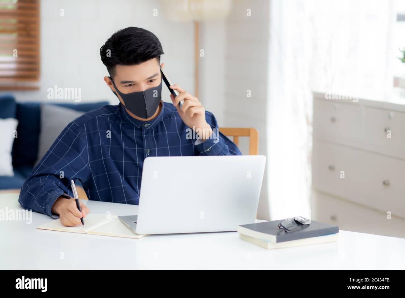 Jeune homme d'affaires asiatique dans le masque de visage parlant smartphone et de travail sur ordinateur portable pour protéger Covid-19, homme d'affaires quarantaine travail de la maison et W Banque D'Images