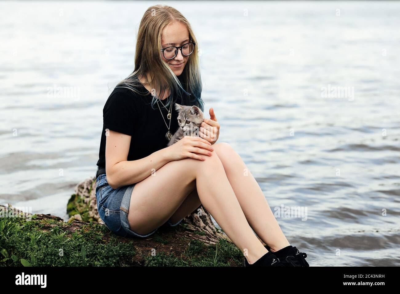 Jeune femme blonde aux cheveux bleus, portant des lunettes, un short en  denim et un t-shirt noir. Assis sur un arbre flottant près du lac avec un  vieux de trois mois Photo