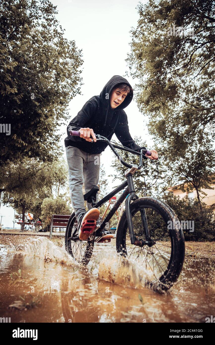 Un adolescent éclabousse de l'eau dans une flaque tout en faisant du vélo pendant la saison des pluies Banque D'Images