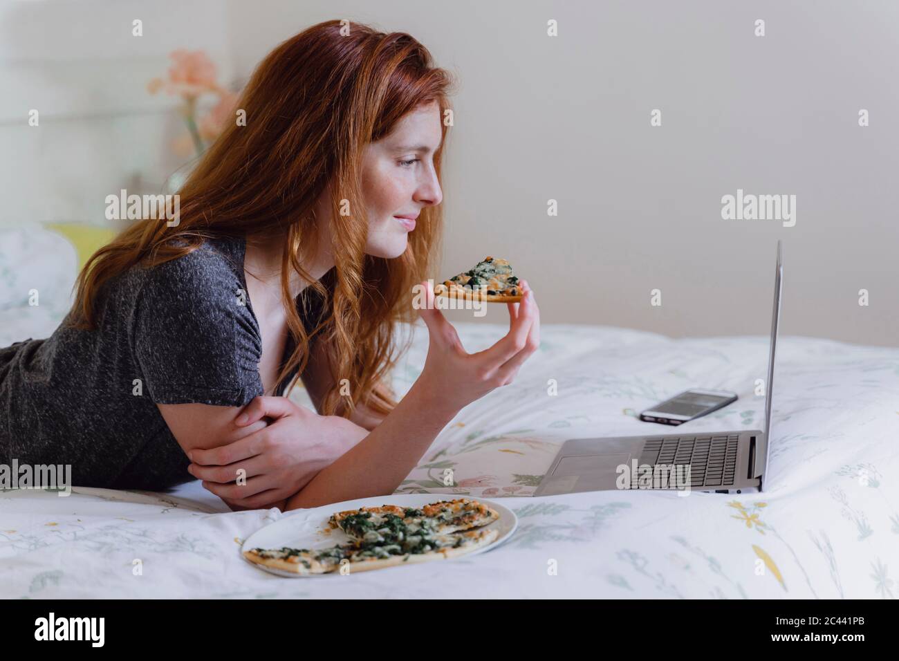 Jeune femme avec de longs cheveux bruns regardant un film sur ordinateur portable tout en appréciant la pizza dans la chambre Banque D'Images