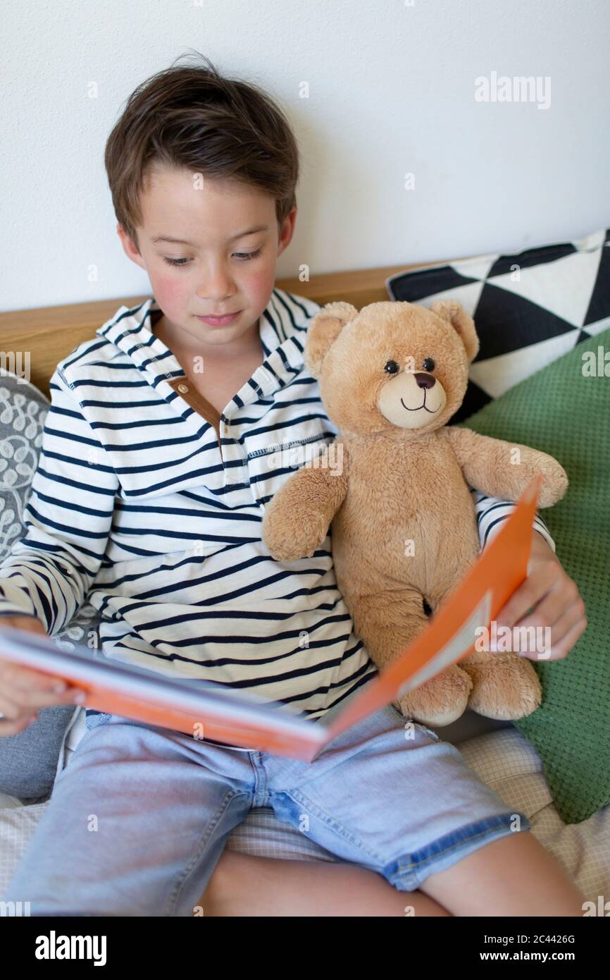 Portrait d'un petit garçon assis sur le lit avec son ours en peluche lisant Banque D'Images