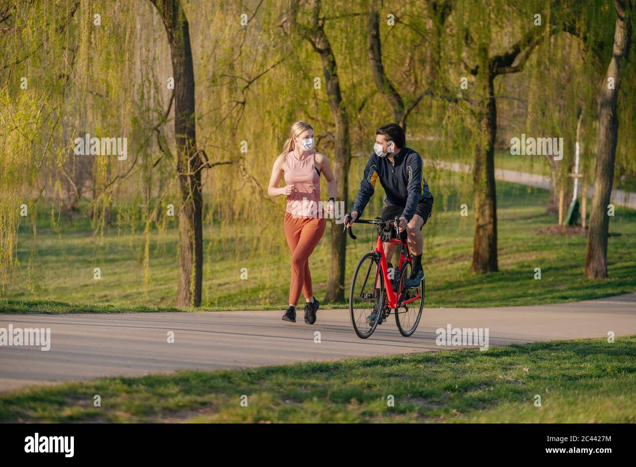 Homme en vélo par femme jogging sur la piste de marche du parc public pendant COVID-19 Banque D'Images