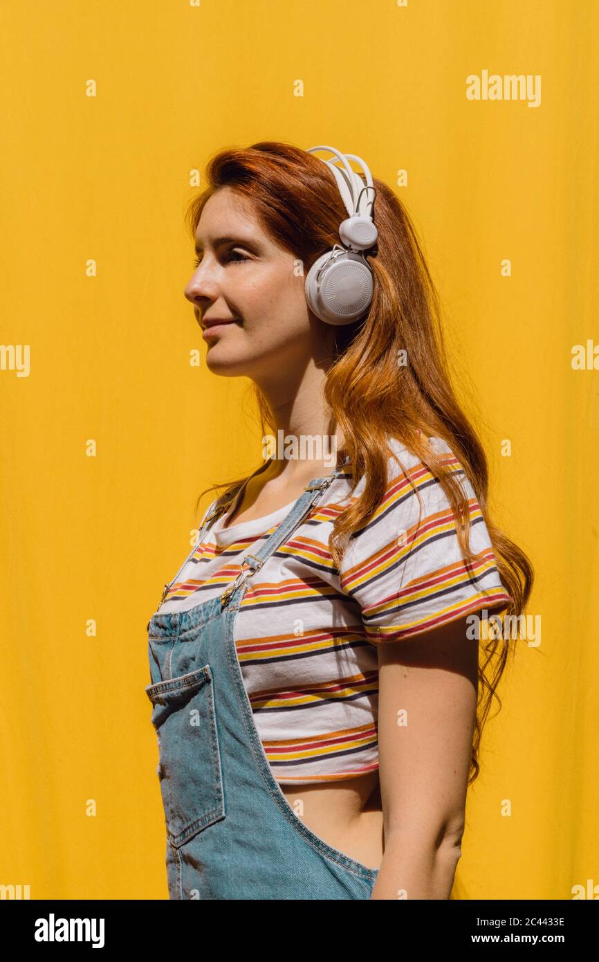 Jeune femme souriante écoutant de la musique avec un casque contre un mur jaune pendant la journée ensoleillée Banque D'Images Jeune femme souriante écoutant de la musique avec un casque contre un mur jaune pendant la journée ensoleillée Banque D'Images