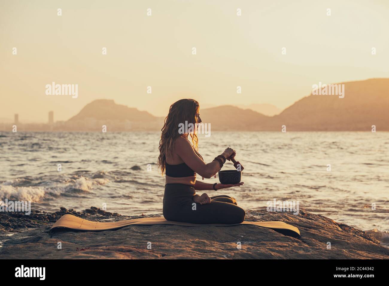 Femme mature méditant avec un bol chantant pratiquant le yoga sur la plage rocheuse en soirée Banque D'Images
