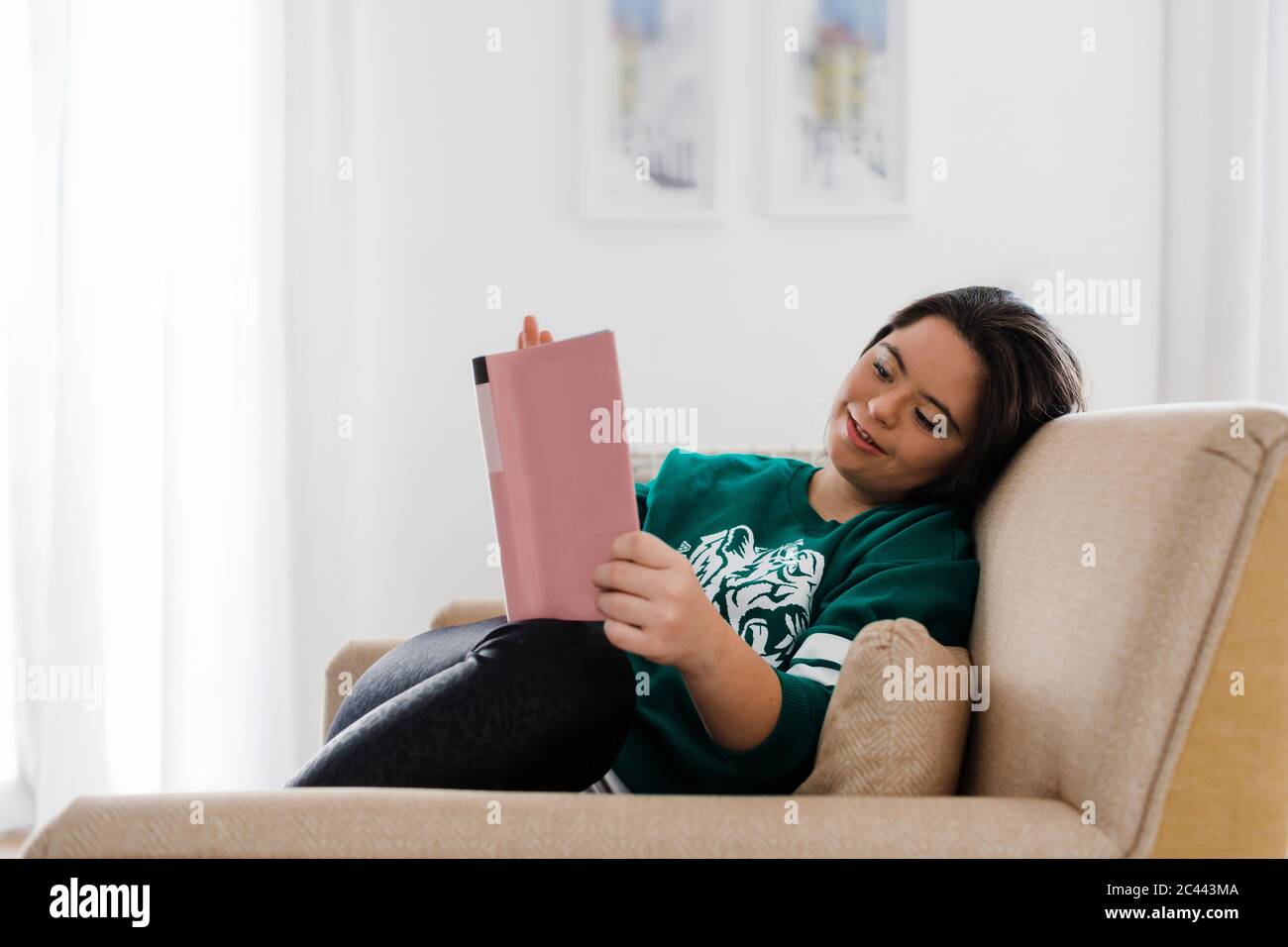 Bonne jeune femme avec le syndrome de Down lire livre tout en se reposant sur un fauteuil à la maison Banque D'Images