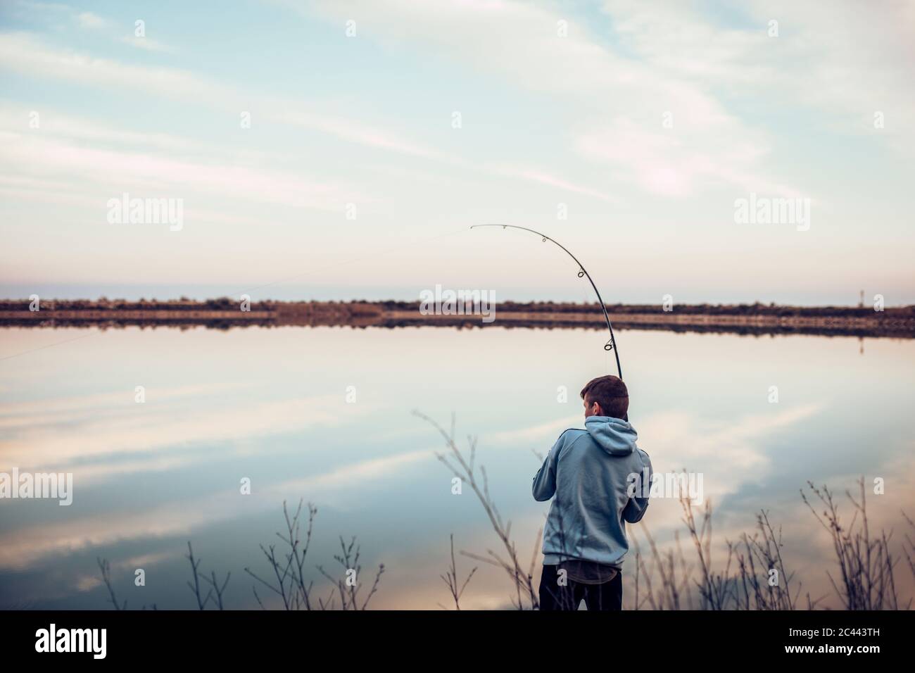 Vue arrière d'un adolescent portant une chemise à capuche, pêche avec la canne dans le lac contre le ciel nuageux Banque D'Images