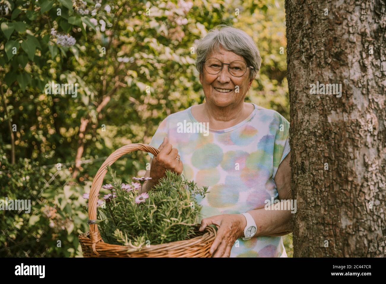 Femme âgée souriante et à la retraite tenant un panier de fleurs et d'herbes tout en se tenant près d'un arbre dans le jardin Banque D'Images