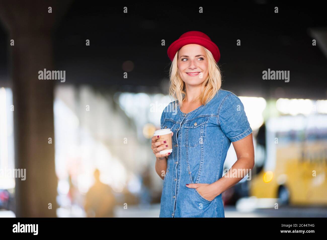 Portrait d'une femme blonde souriante avec café à emporter, portant une robe en denim et un chapeau rouge Banque D'Images