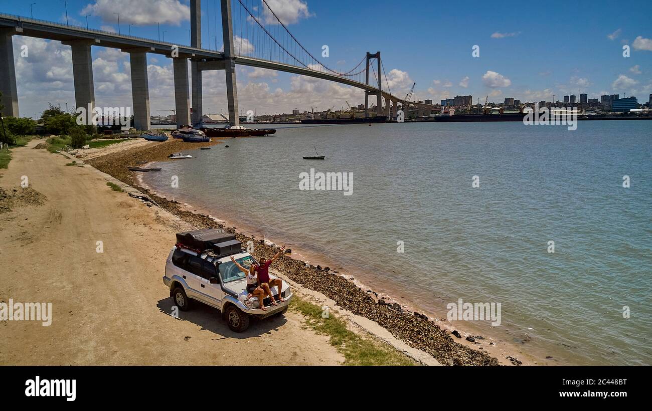 Mozambique, Katembe, couple adulte assis sur le capot d'une voiture 4x4 et agitant à la caméra avec la ville et le pont Maputo-Katembe en arrière-plan Banque D'Images