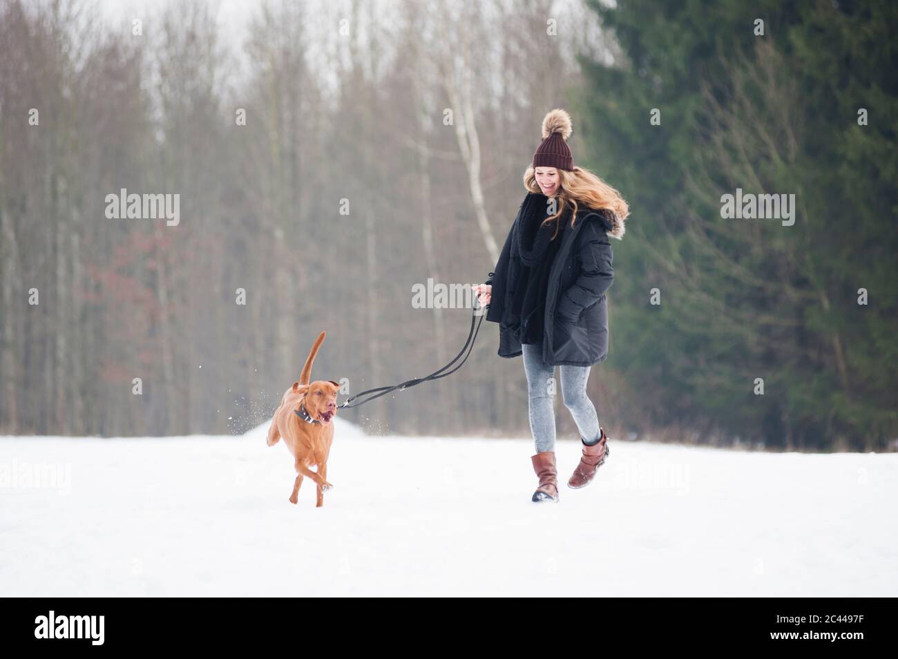 Bonne jeune femme qui court avec un chien sur la neige contre les arbres en forêt en hiver Banque D'Images