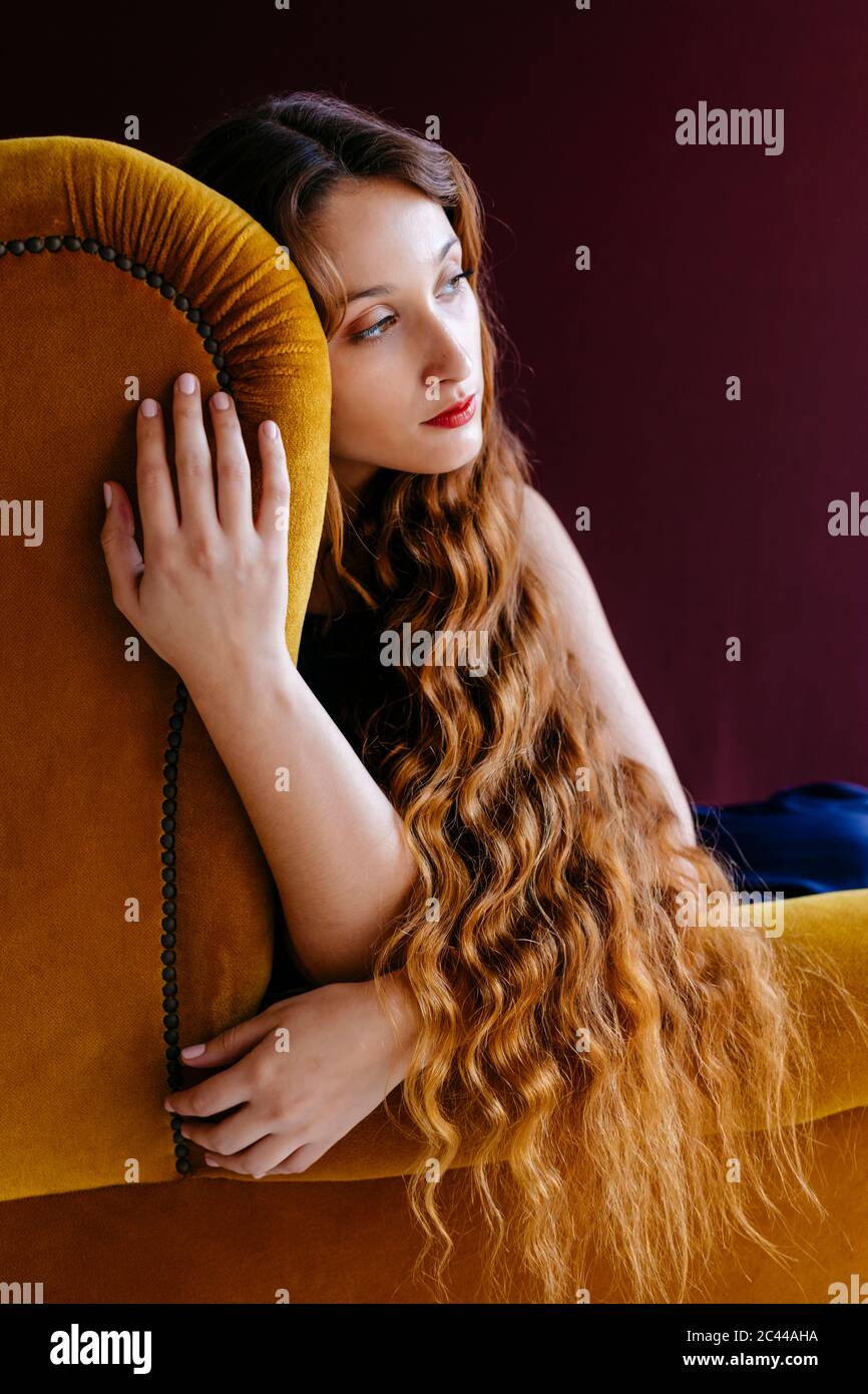 Jeune femme attentionnés avec de longs cheveux ondulés bruns assis sur une chaise dorée sur un fond coloré Banque D'Images