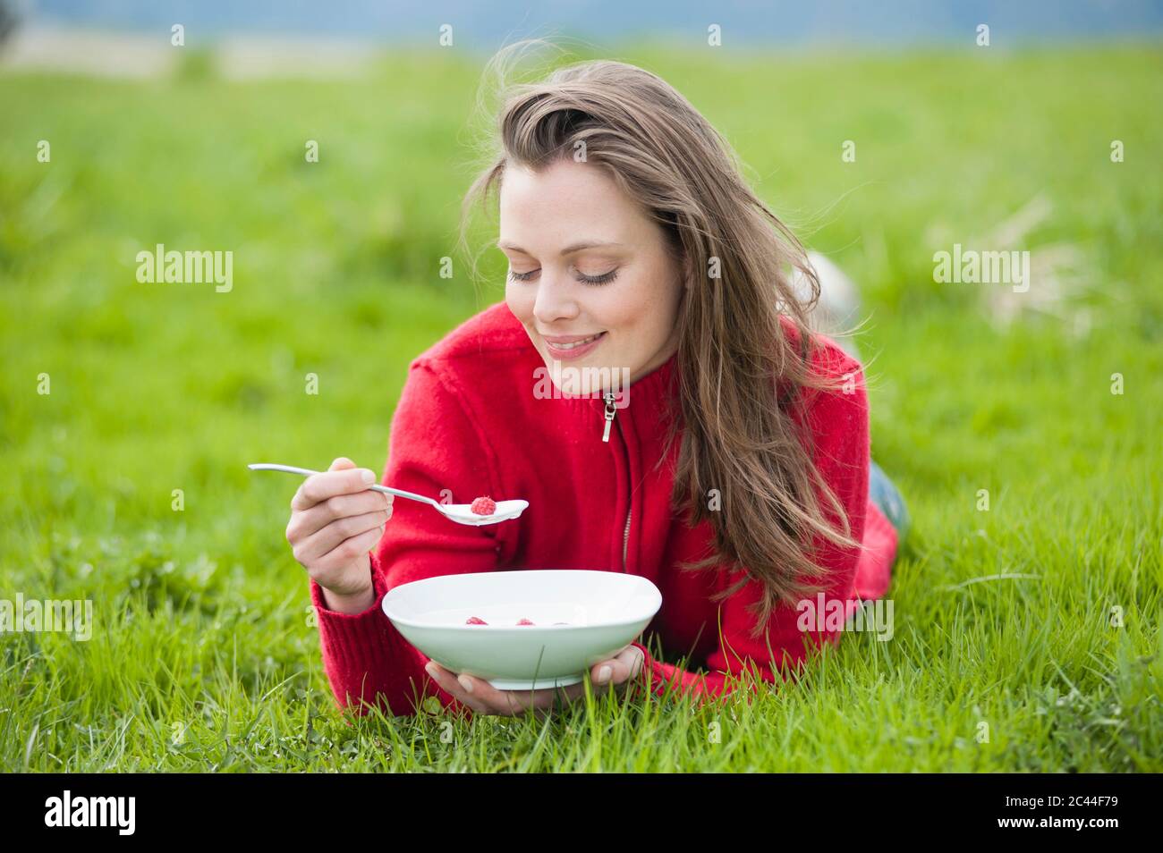 Souriante belle femme mangeant du yaourt frais et de la framboise tout en étant allongé sur l'herbe Banque D'Images