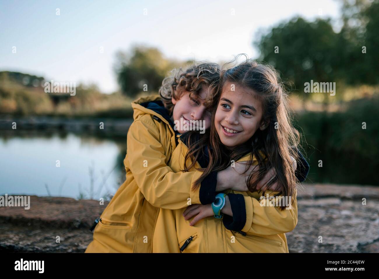 Garçon embrassant une sœur souriante portant des manteaux de pluie jaunes sur la rive Banque D'Images