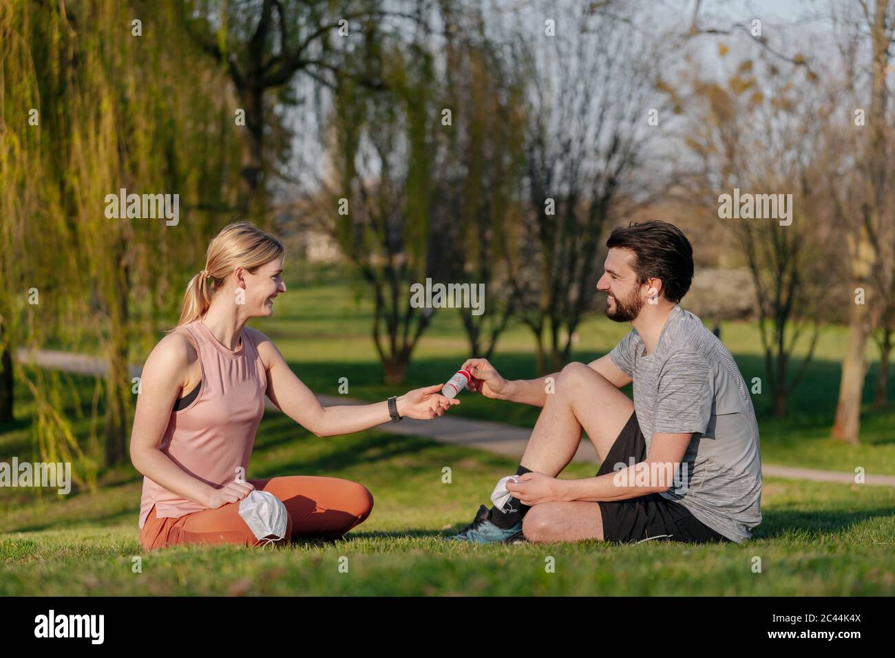 Homme souriant donnant de l'assainisseur pour les mains à une femme assise sur l'herbe au parc Banque D'Images