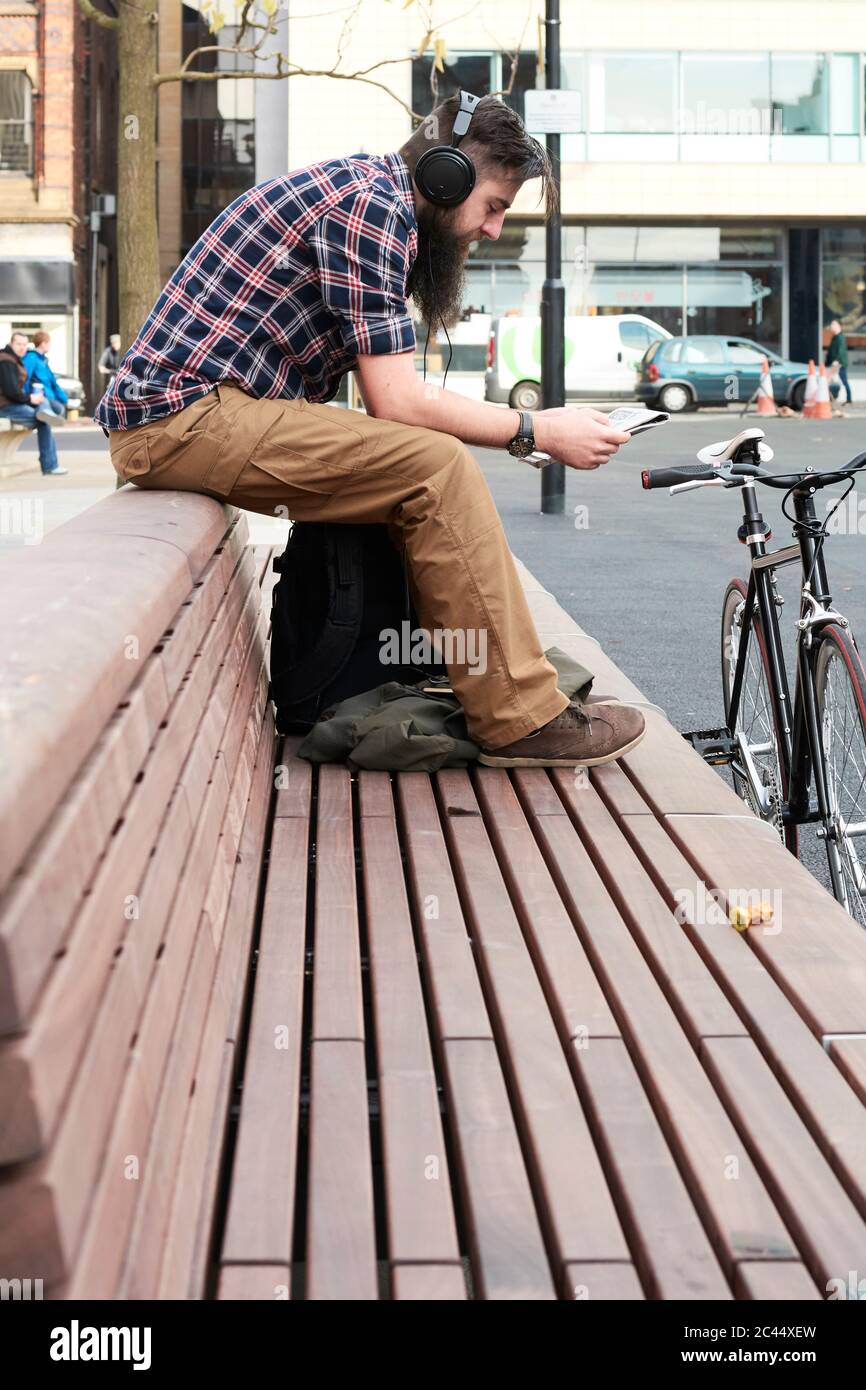 Vue latérale du bearded man reading newspaper while sitting on bench in city Banque D'Images
