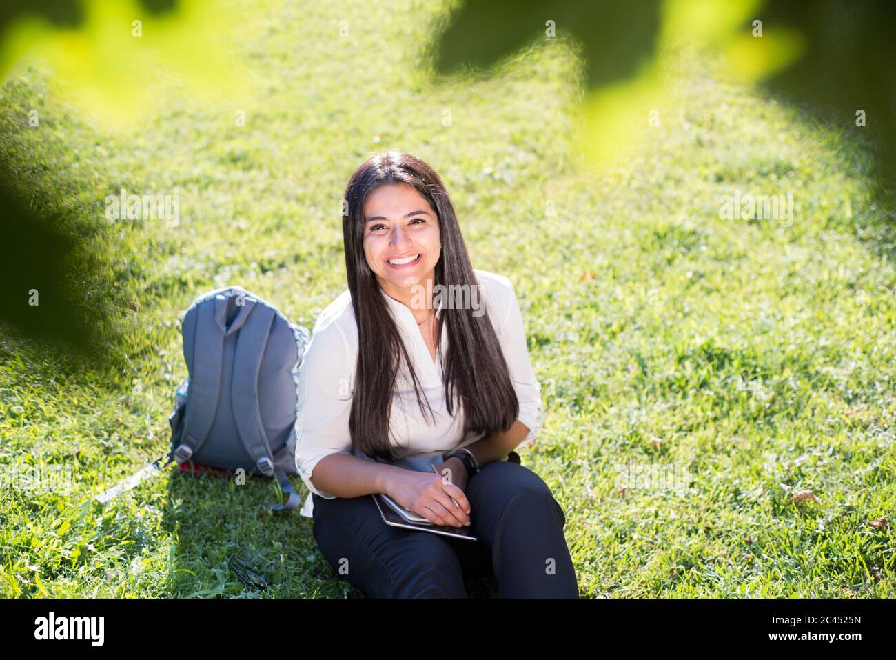 Une fille indienne, étudiante, sourit, s'assoit sur l'herbe verte, avec un sac à dos et un bloc-notes, écrit Banque D'Images