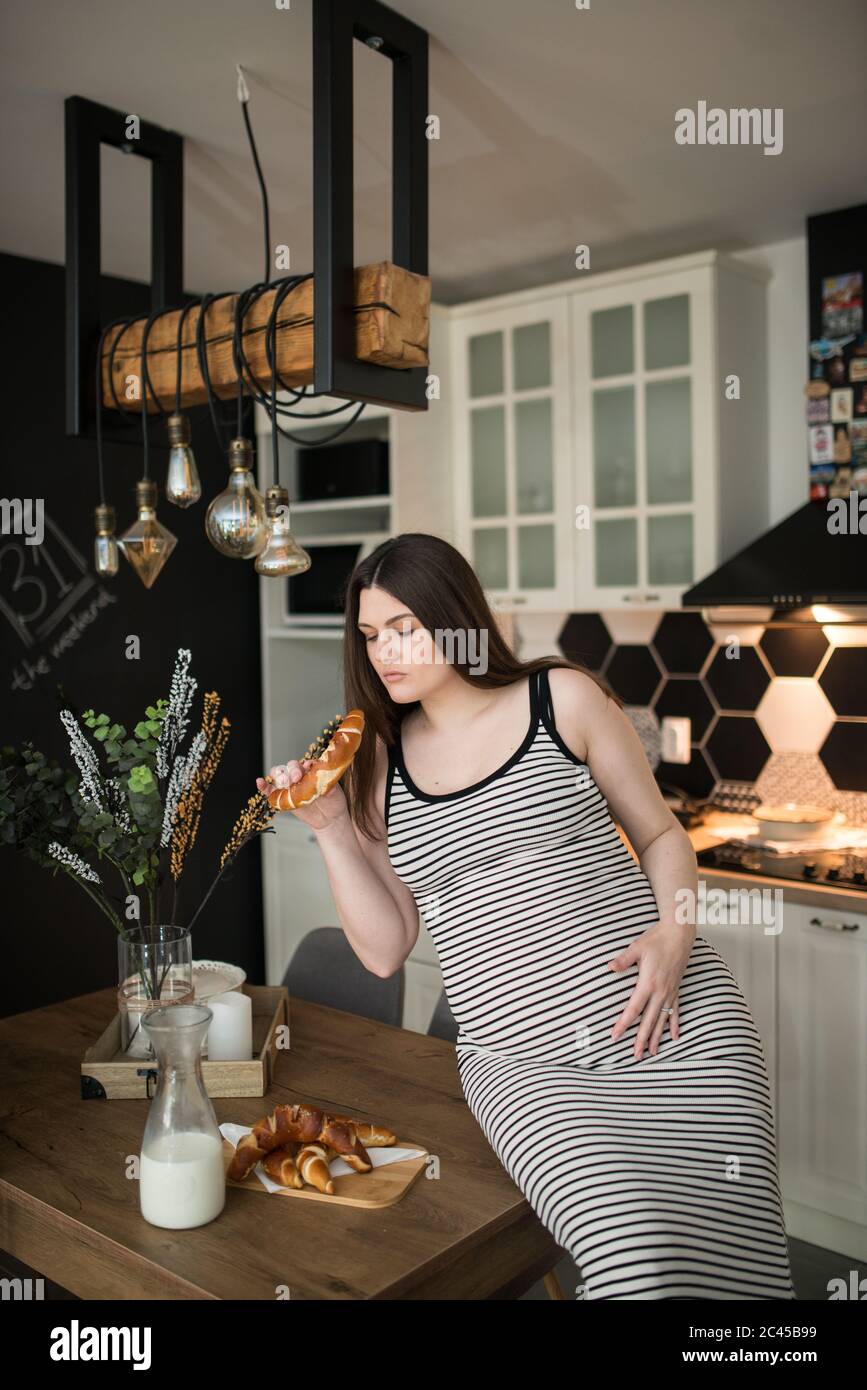 une jeune femme enceinte dans sa cuisine mangeant un muffin. Une femme enceinte dans la cuisine s'assoit à une table et mange Banque D'Images