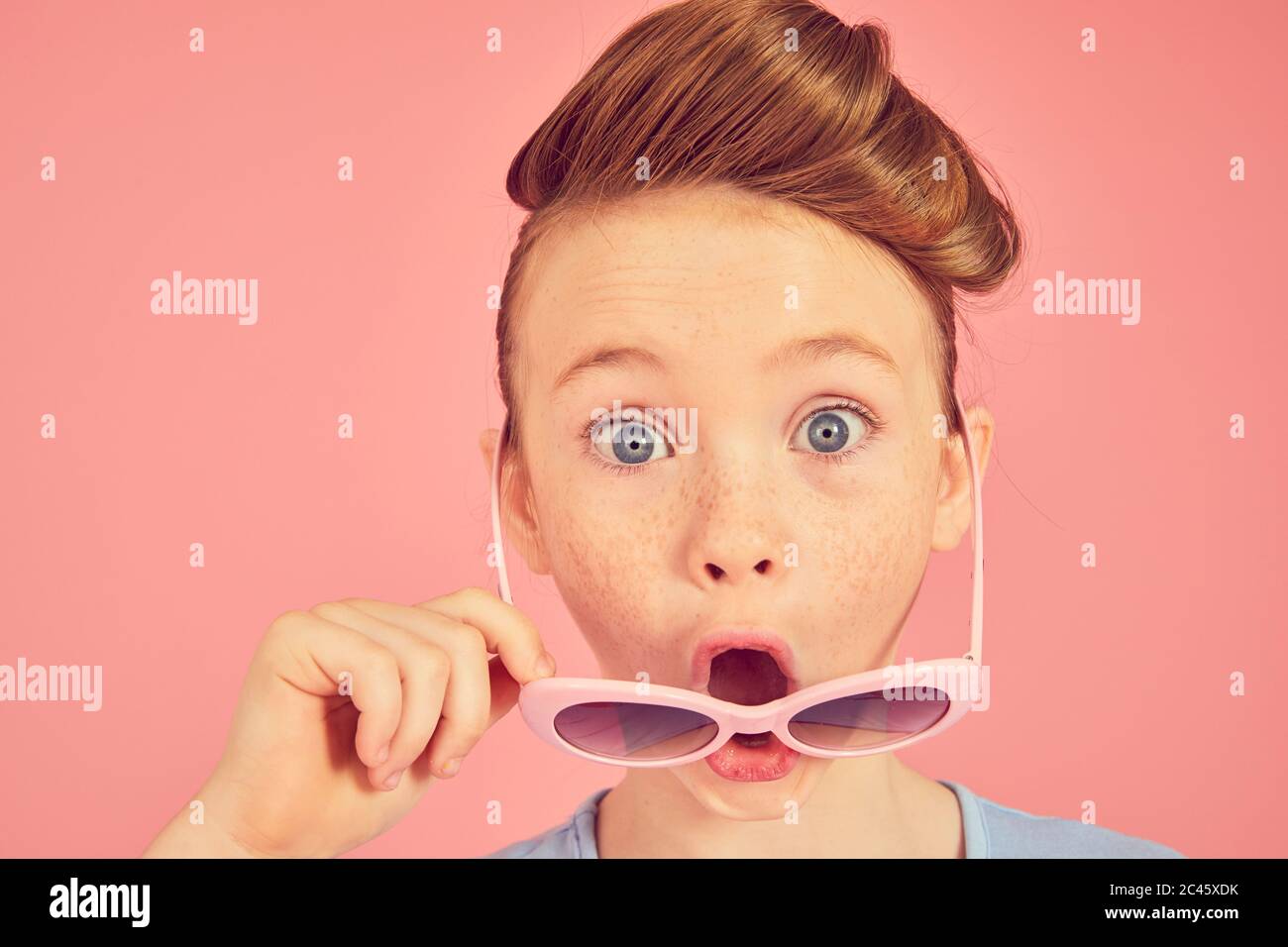 Portrait de la fille de brunette sur fond rose, regardant l'appareil photo au-dessus de ses lunettes de soleil, bouche ouverte. Banque D'Images