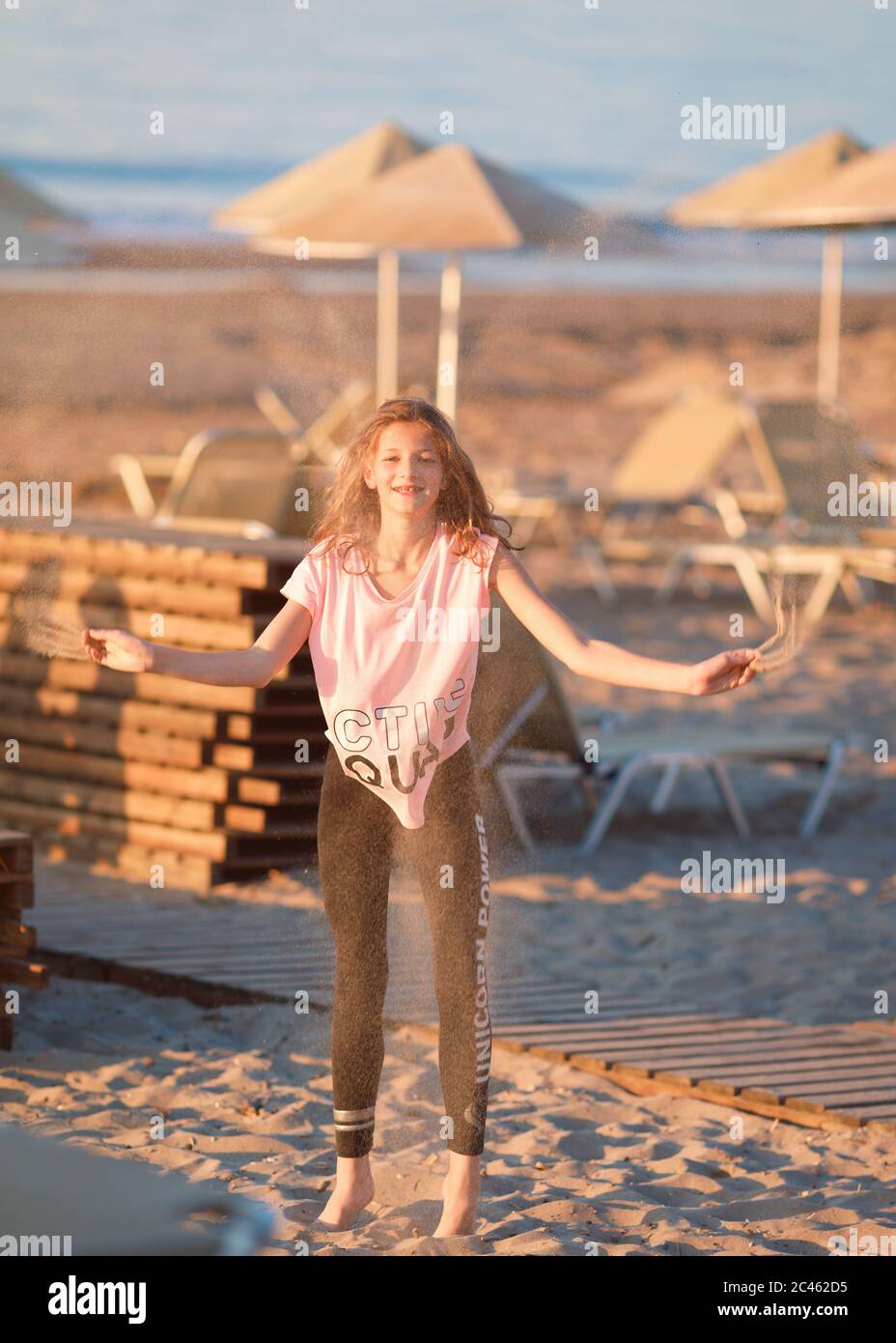 Jeune fille, entre les âges, portant des vêtements décontractés d'été, jetant du sable dans les airs sur la plage Banque D'Images