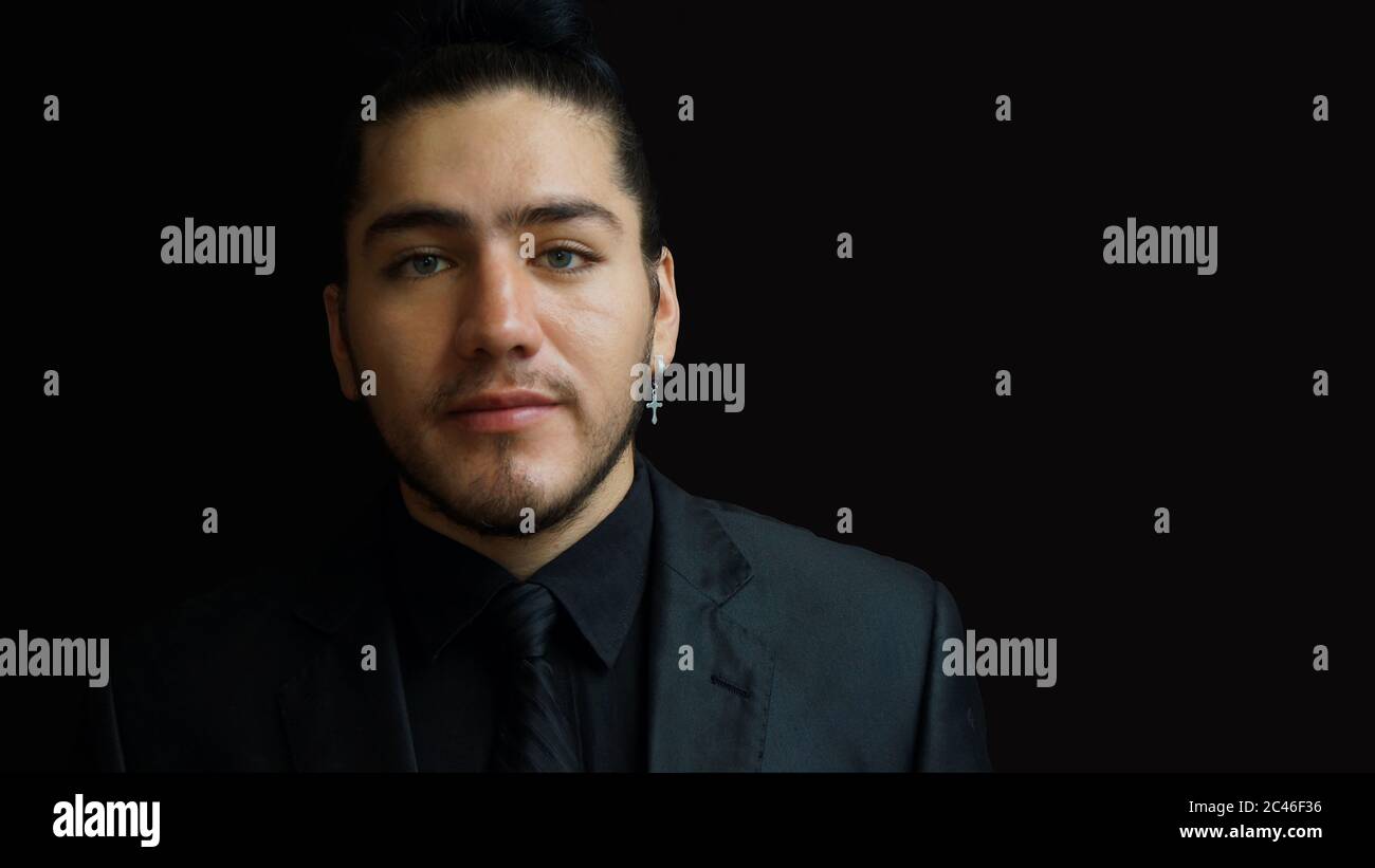Portrait d'un jeune homme latin portant un contour d'oreille avec un costume noir, une chemise noire et une cravate noire sur fond noir Banque D'Images