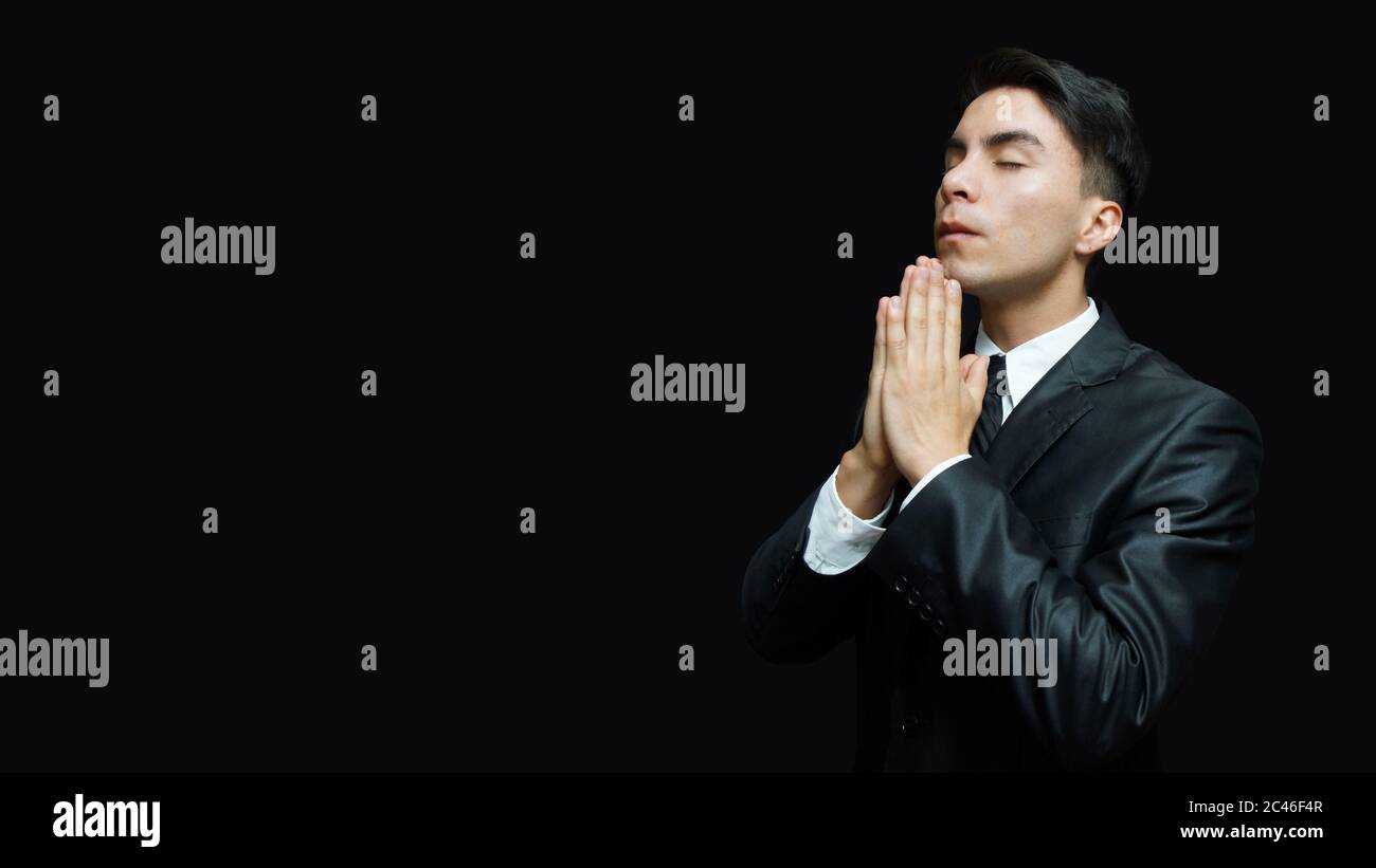 Jeune latino en costume noir, chemise blanche, cravate noire avec ses mains en position de prière sur fond noir Banque D'Images