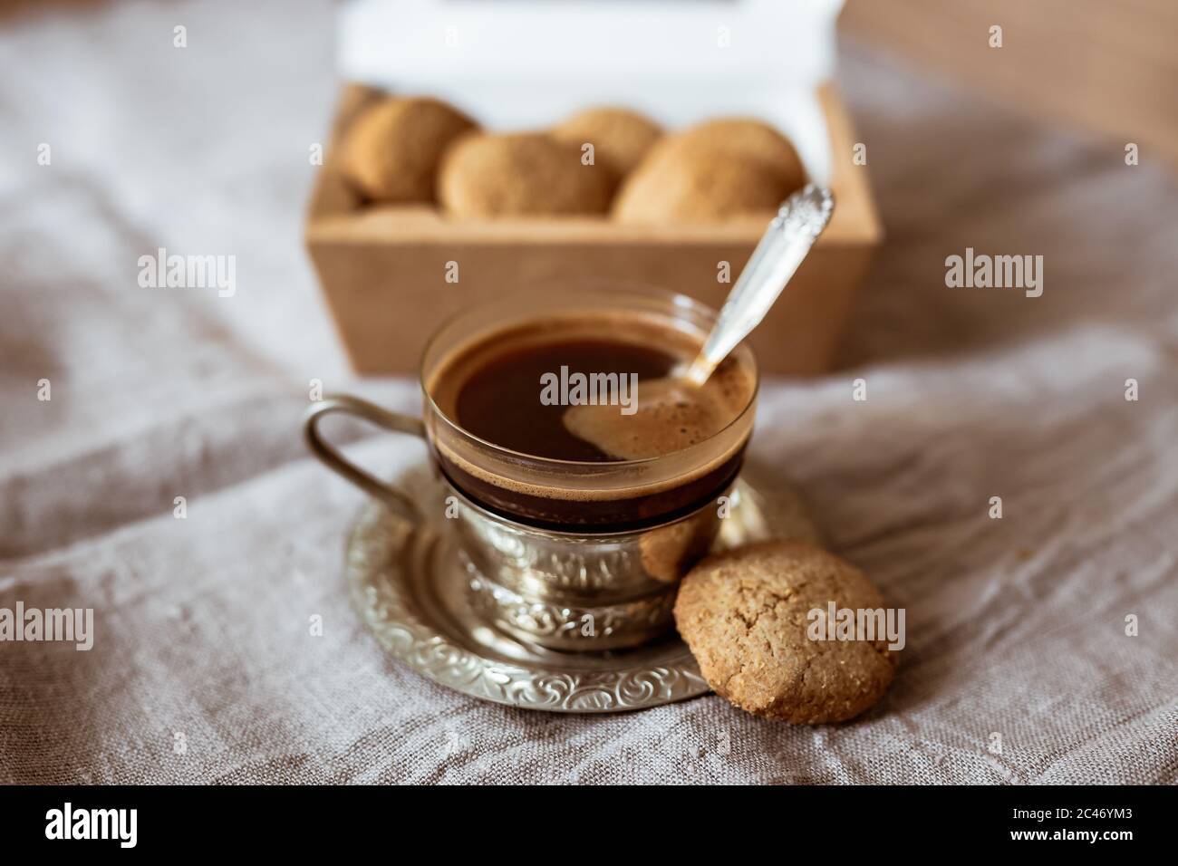 Une photo en gros plan d'une belle tasse de café noir en verre dans un porte-verre sur une soucoupe en métal et des biscuits dans une boîte en carton Banque D'Images