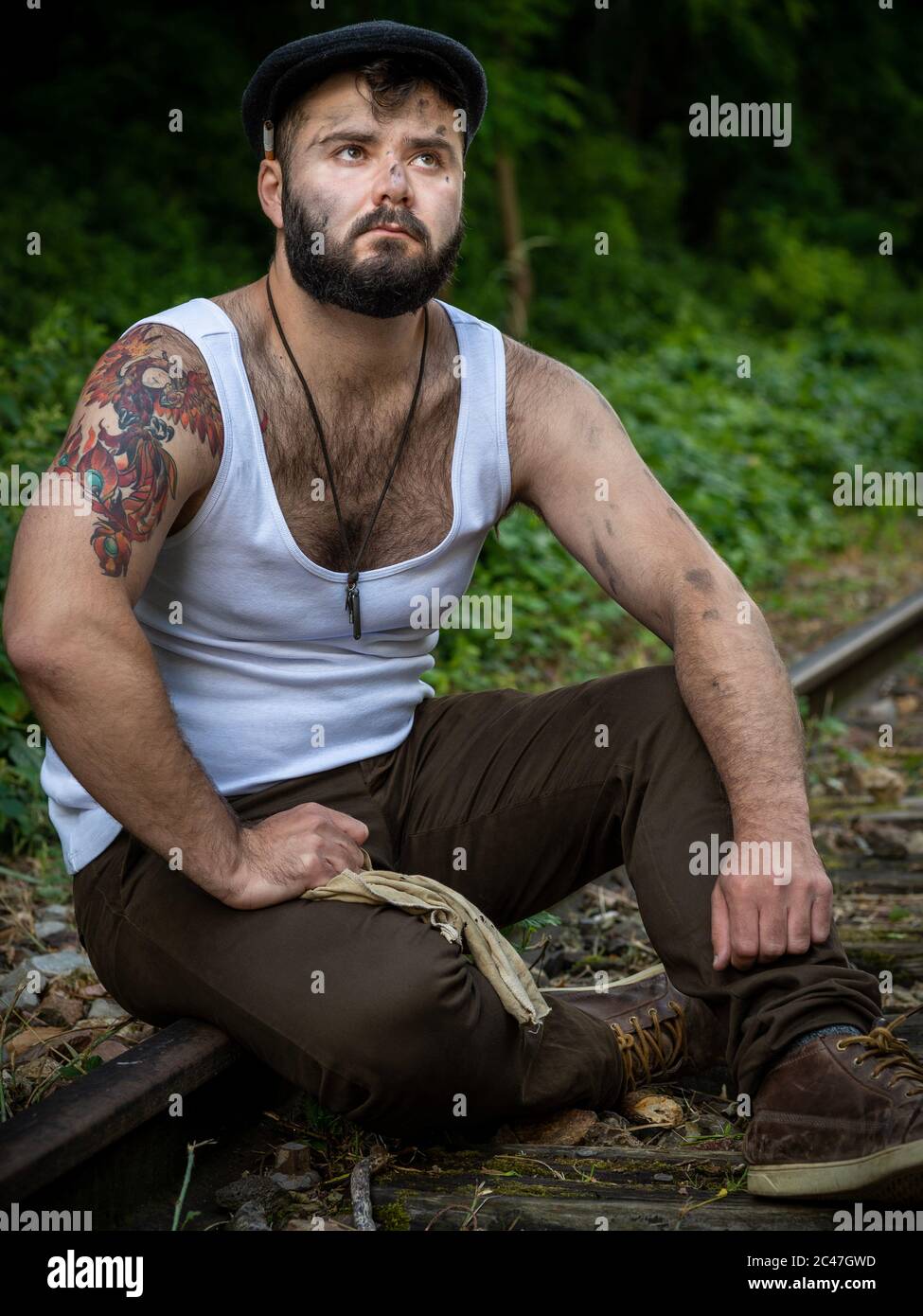 Jeune homme barbu français, avec des tatouages et des taches de charbon de bois sur sa peau, pris à l'extérieur sur une voie ferrée désutilisée avec lumière naturelle à Paris, France Banque D'Images