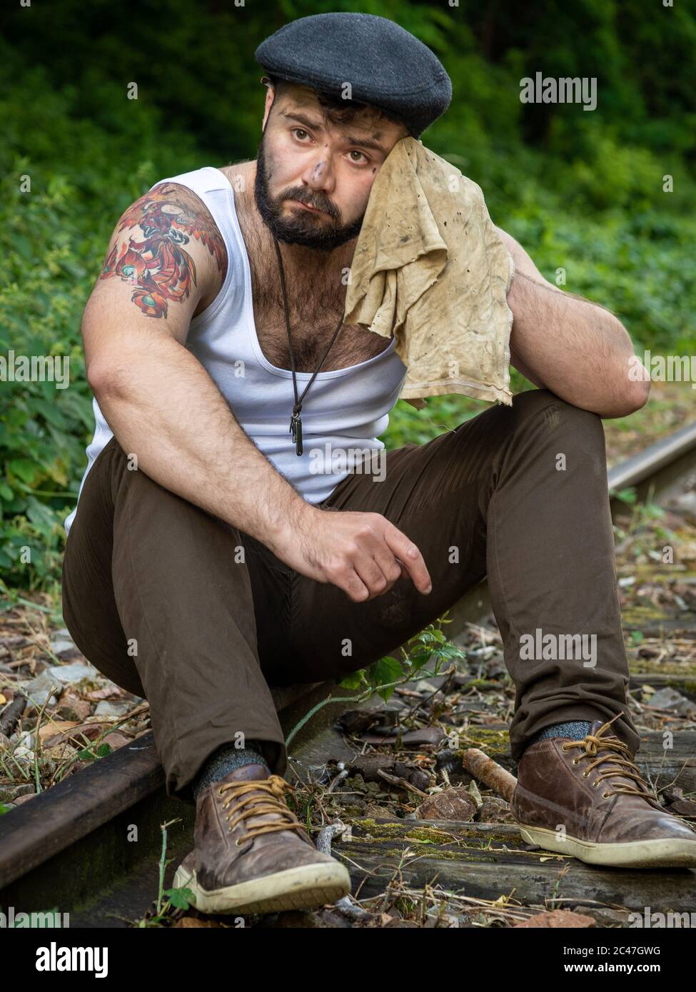 Jeune homme barbu français, avec des tatouages et des taches de charbon de bois sur sa peau, pris à l'extérieur sur une voie ferrée désutilisée avec lumière naturelle à Paris, France Banque D'Images