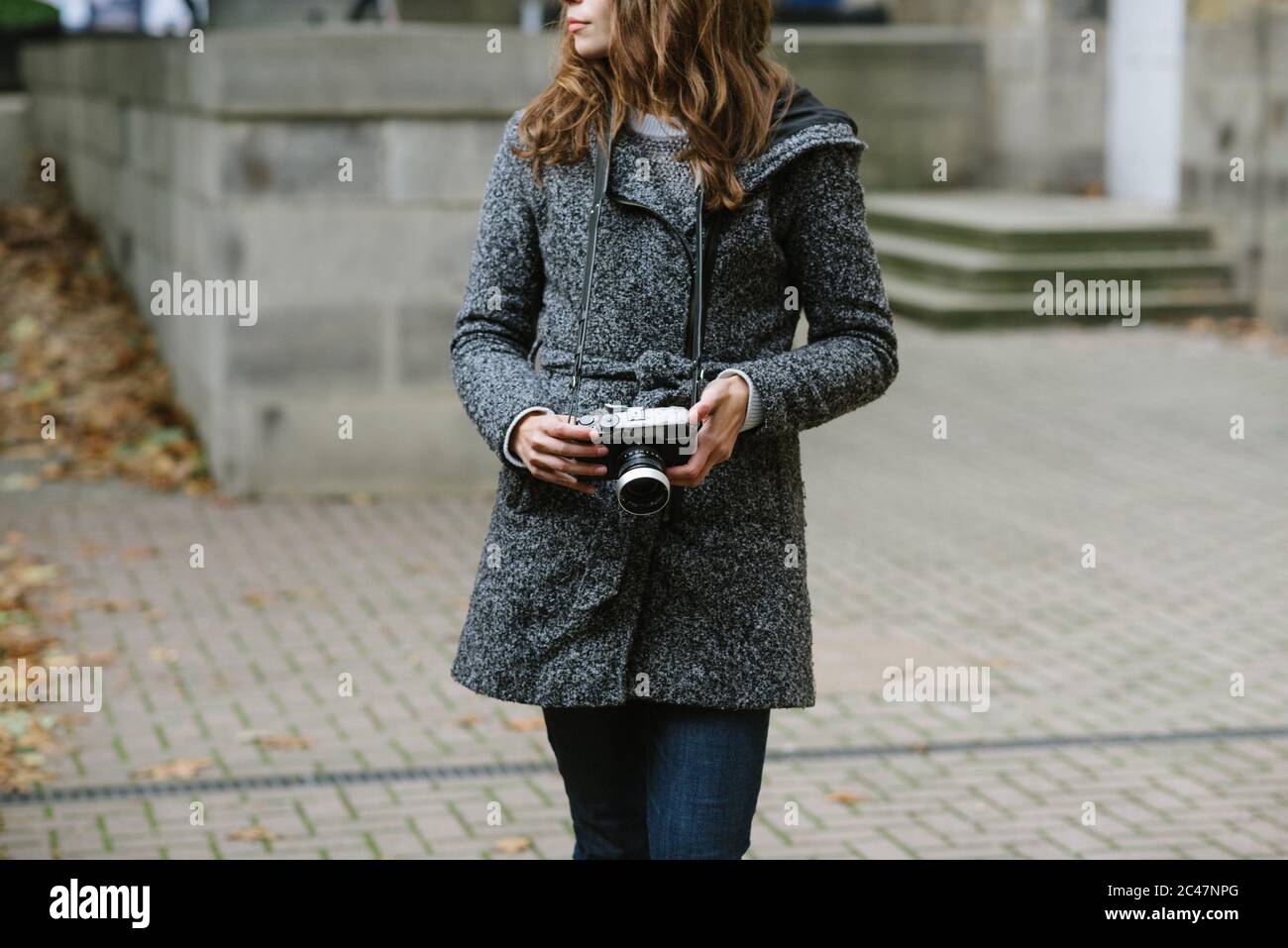 Belle femme debout à l'extérieur, portant un Jean bleu et un manteau gris et tenant un appareil photo vintage Banque D'Images