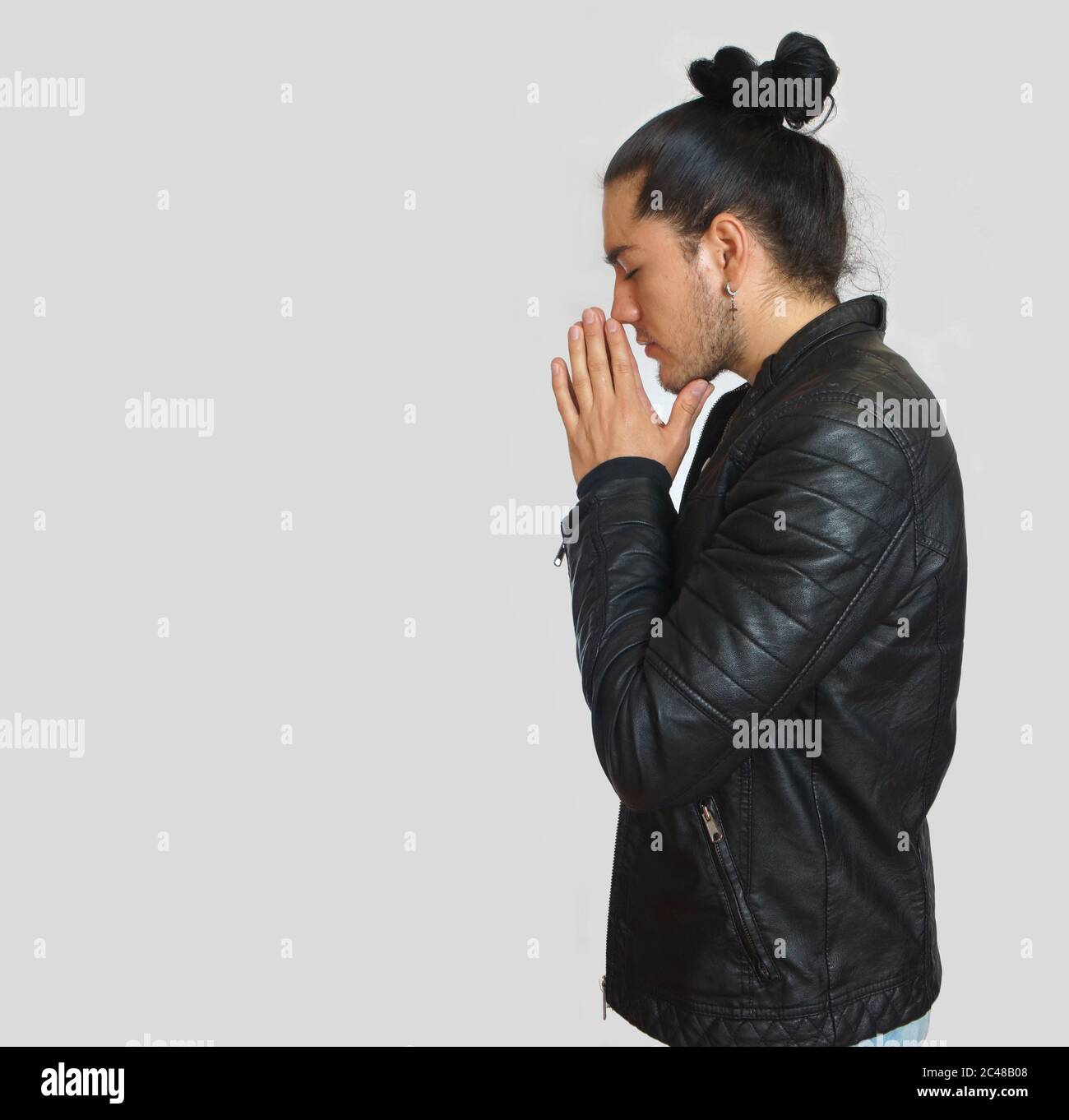 Jeune hispanique avec les cheveux rassemblés fait noeud portant le t-shirt noir et la veste en cuir noir, avec ses mains jointes en position de prière avec crouppé Banque D'Images
