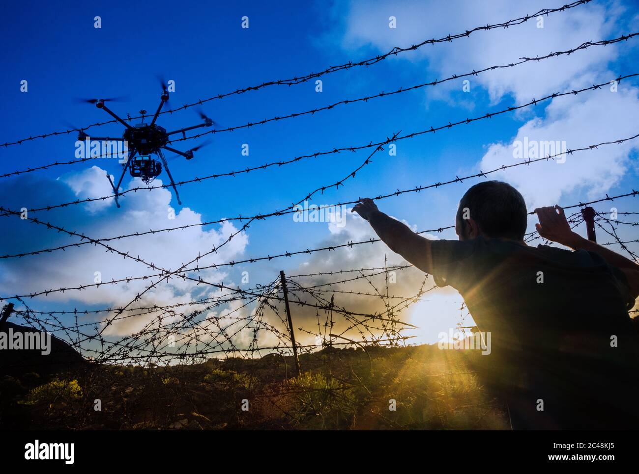 Homme regardant à travers la clôture barbelée au lever du soleil avec un drone au-dessus. Brexit, immigration, asile, contrebande de personnes, contrôle des frontières... concept. Banque D'Images