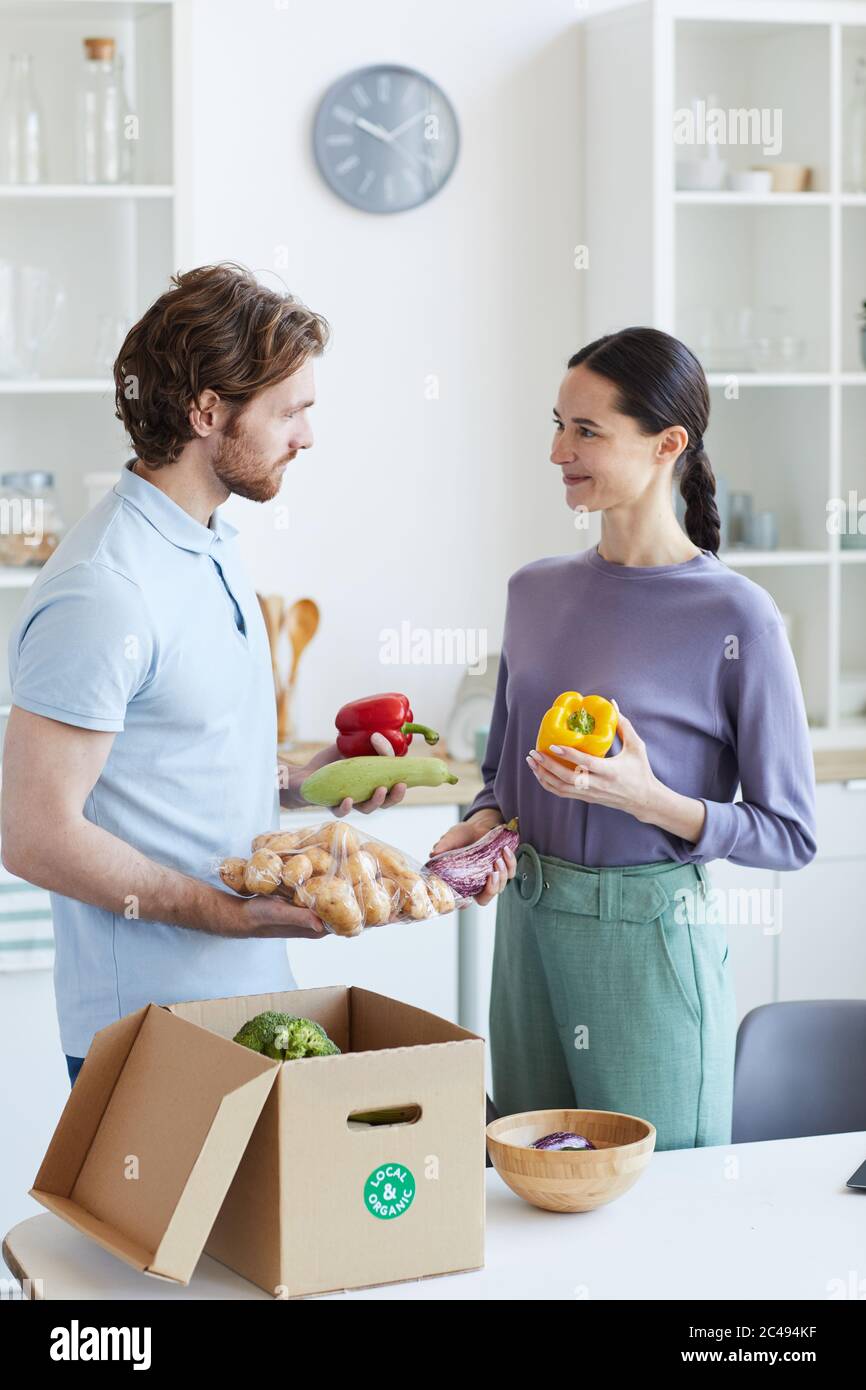 Jeune couple prenant des légumes frais hors de la boîte et parlant les uns avec les autres en se tenant dans la cuisine Banque D'Images