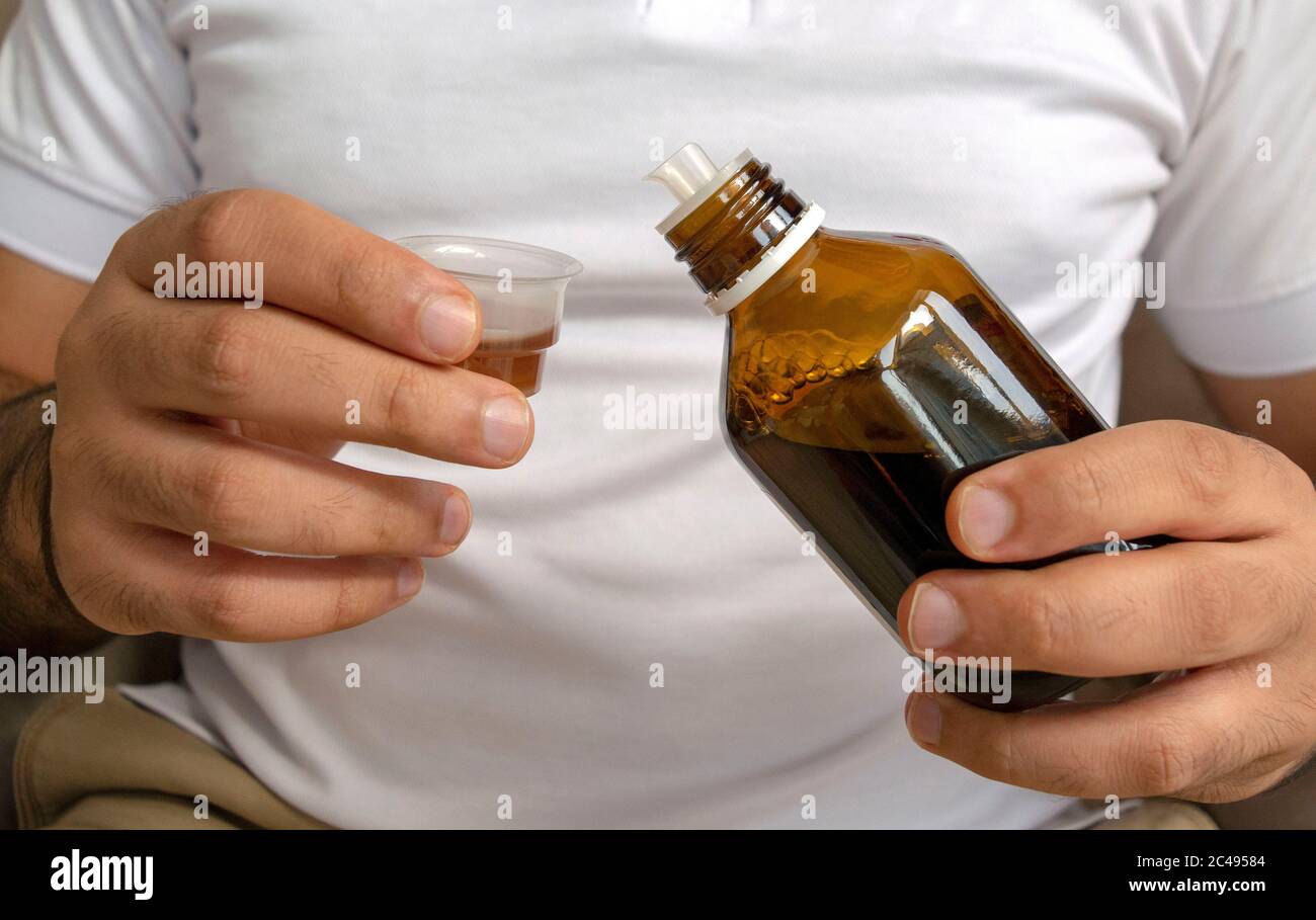 Jeune homme en t-shirt blanc verse du sirop médical dans une tasse à mesurer. Le jeune homme tient une bouteille de sirop avec une tasse. Banque D'Images
