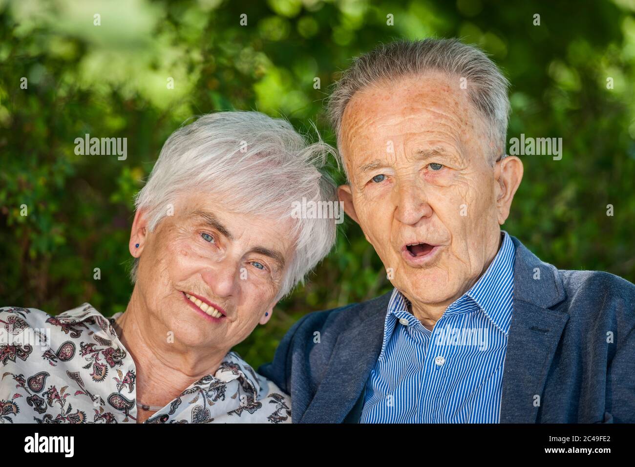 Portrait tête-et-épaule d'un couple de retraités âgés devant des feuilles vertes en été regardant avec joie la caméra. Homme avec la bouche ouverte. Banque D'Images