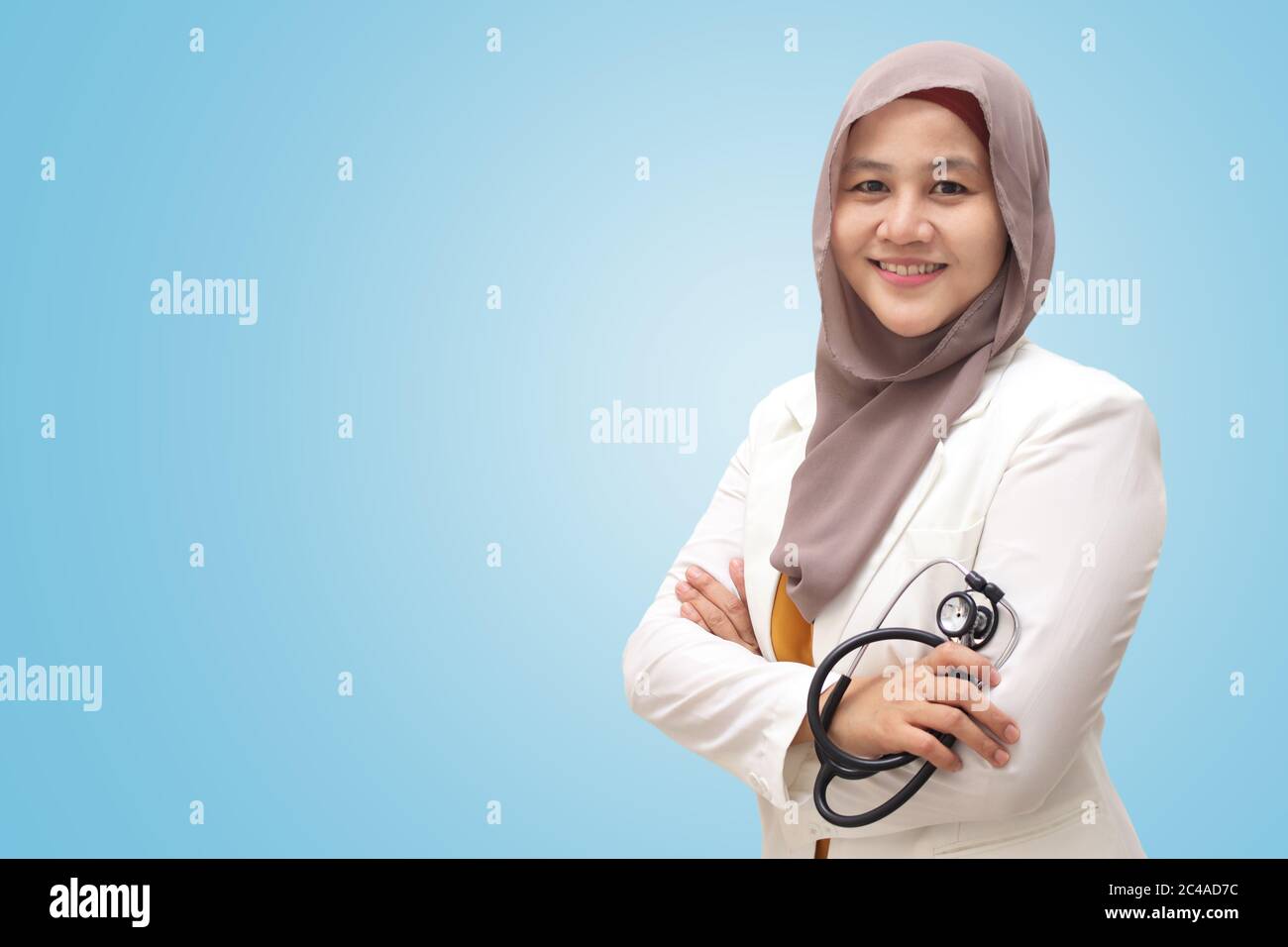 Femme musulmane médecin portant le hijab et la combinaison, debout avec bras croisés et tenant le stéthoscope, personne réussie et confiante, sur fond bleu Banque D'Images