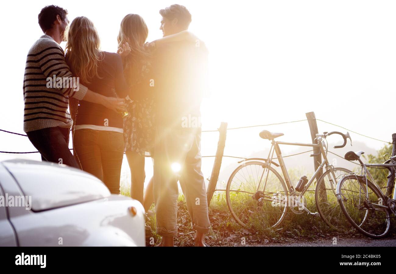 Quatre personnes se tenant sur le côté d'une route, voiture garée et vélos se penchant contre une clôture, lumière du soleil. Banque D'Images