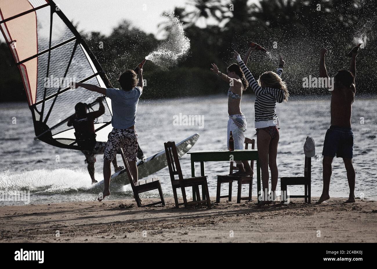 Petit groupe d'hommes et de femmes se tenant sur une plage de sable autour d'une table et de chaises, bras levés, tenant des bouteilles, regardant un coupe-vent. Banque D'Images