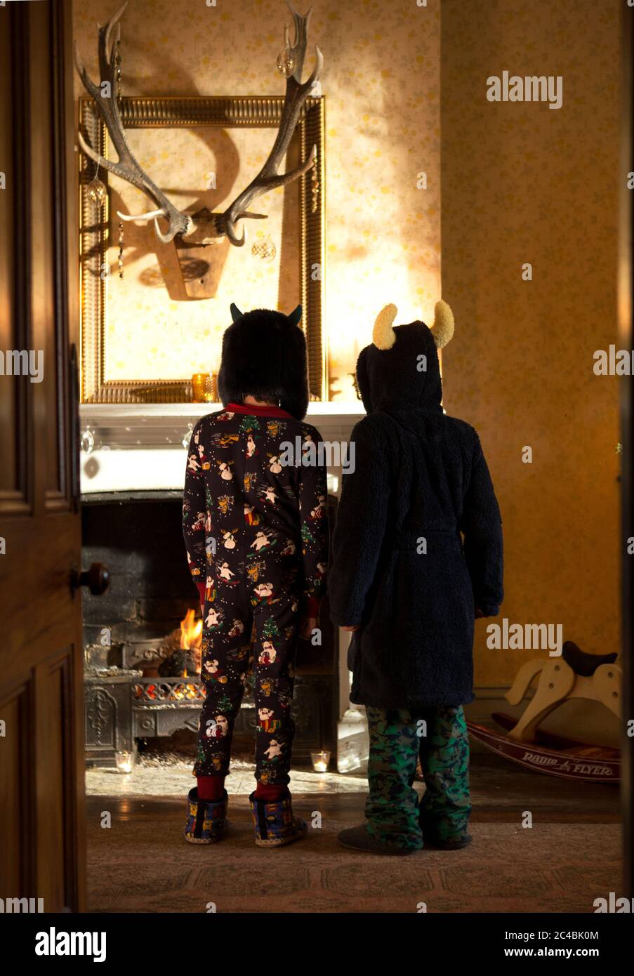 Vue arrière de deux enfants portant un pyjama, un peignoir en éponge et des chapeaux devant une cheminée, trophée de chasse sur le mur. Banque D'Images