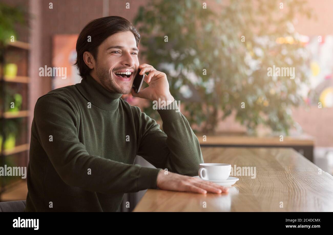 Un gars enthousiaste qui parle sur son smartphone et qui boit du café à la cafétéria Banque D'Images