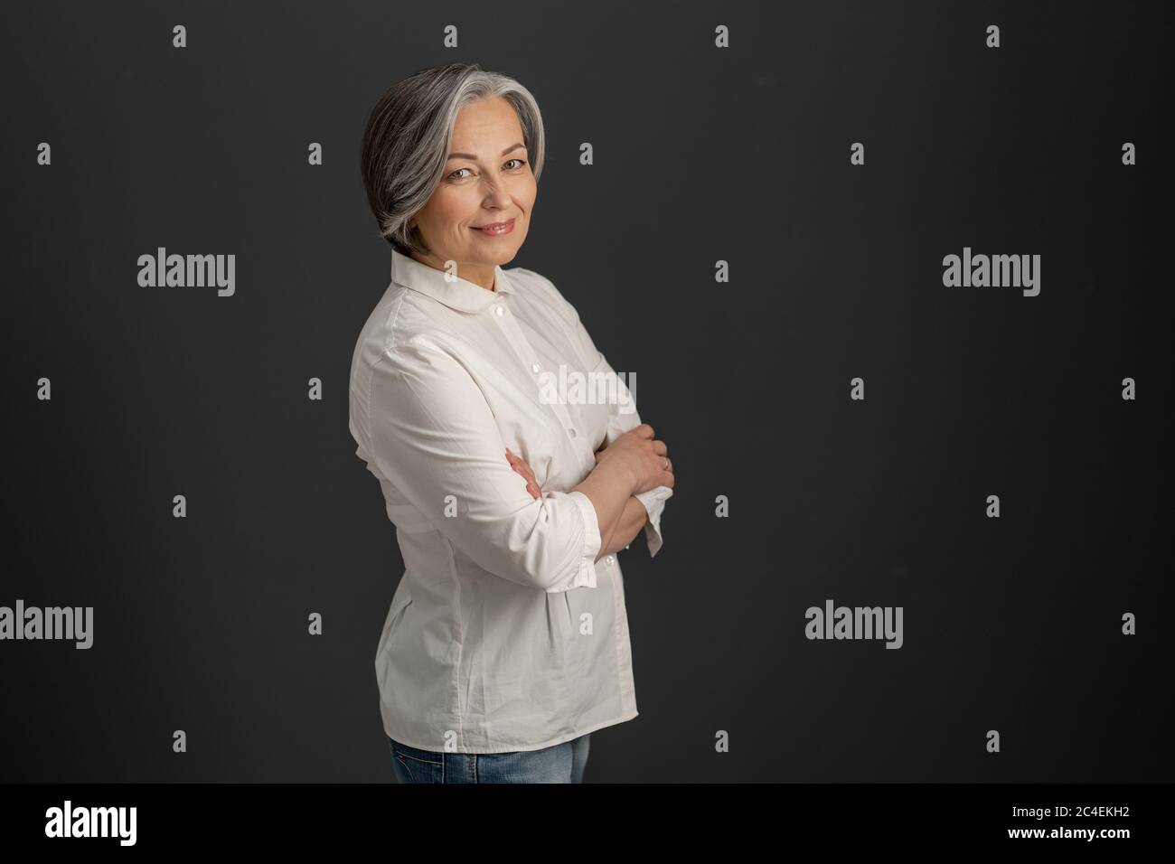 Une jolie femme aux cheveux blancs sourit en regardant l'appareil photo avec les bras croisés. Femme d'affaires caucasienne élégante isolée sur fond gris foncé. Espace texte à Banque D'Images