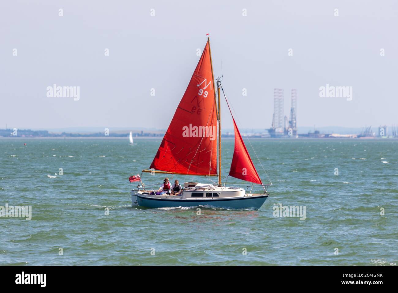 Southend-on-Sea, Royaume-Uni. 26 juin 2020. Un petit voilier avec des voiles rouges marquées 'M 39' glisse à travers l'estuaire de la Tamise près de Chalkwell Beach, Southend. Deux personnes sont à bord, et un drapeau rouge flotte à la poupe. Au loin, une structure industrielle s’élève au-delà de la ligne de flottaison, contrastant loisirs et infrastructures. Penelope Barritt/Alamy Live News Banque D'Images