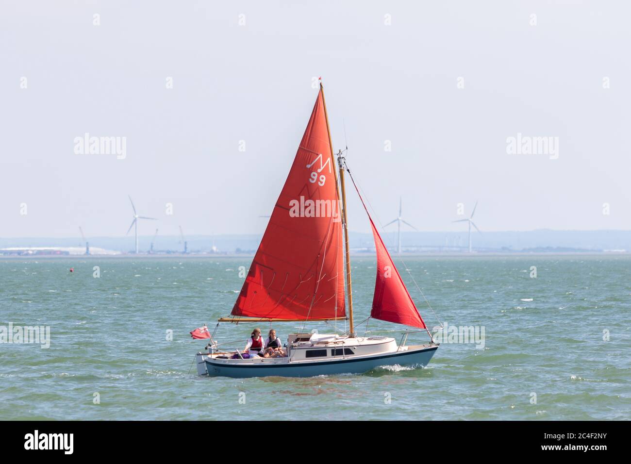 Southend-on-Sea, Royaume-Uni. 26 juin 2020. Un petit voilier avec des voiles rouges marquées 'M 39' glisse à travers l'estuaire de la Tamise près de Chalkwell Beach, Southend. Deux personnes sont à bord, et un drapeau rouge flotte à la poupe. Au loin, une structure industrielle s’élève au-delà de la ligne de flottaison, contrastant loisirs et infrastructures. Penelope Barritt/Alamy Live News Banque D'Images