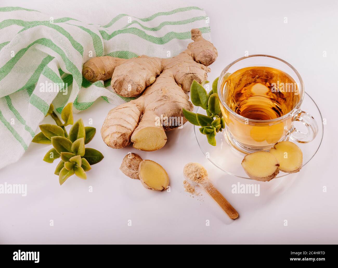 Une tasse de thé de gingembre en verre avec une racine de gingembre frais à côté et quelques feuilles vertes autour, il y a un tissu blanc et vert au coin. Banque D'Images