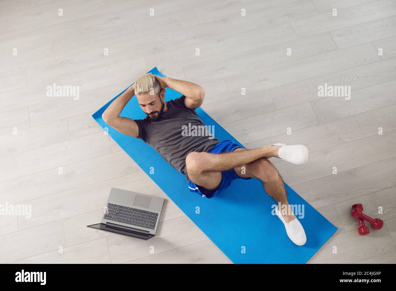 Un jeune homme en forme fait des exercices d'entraînement sur l'abs tout en étant allongé sur le sol, regarde un cours d'entraîneur en ligne tout en ayant un ordinateur portable dans la chambre. Banque D'Images