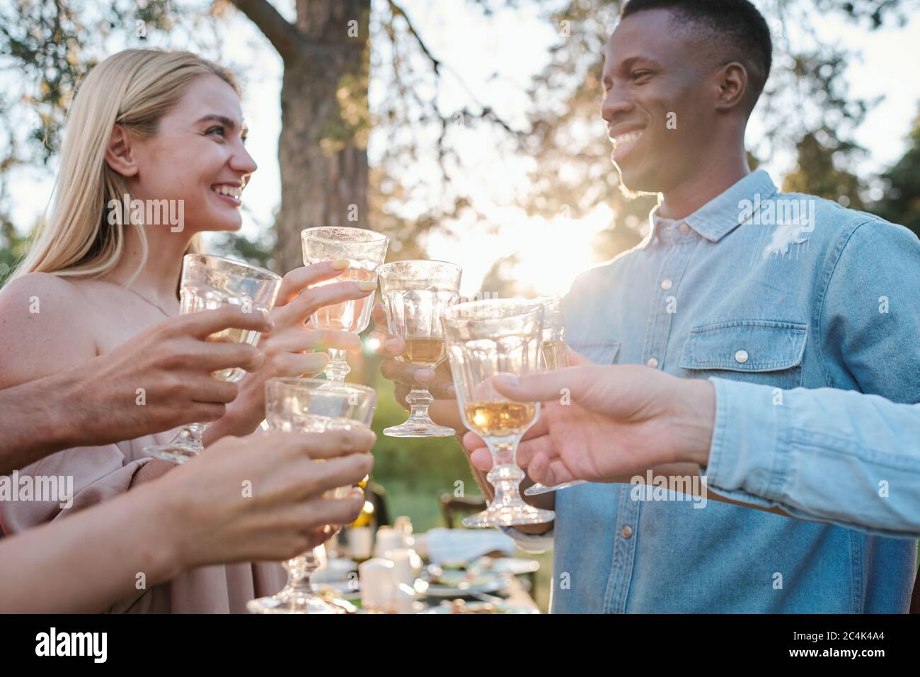 Joyeux jeune interculturel dates regardant les uns les autres avec des sourires torontes tout en se clinquant avec des verres de vin parmi leurs amis à l'extérieur Banque D'Images