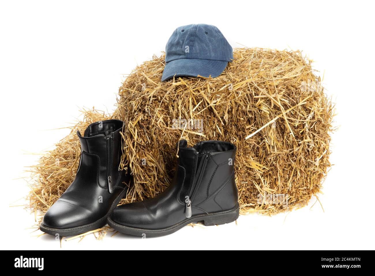mode agricole, chapeau en denim bleu et bottes en cuir noir sur une balle de paille dans une grange isolée sur blanc Banque D'Images