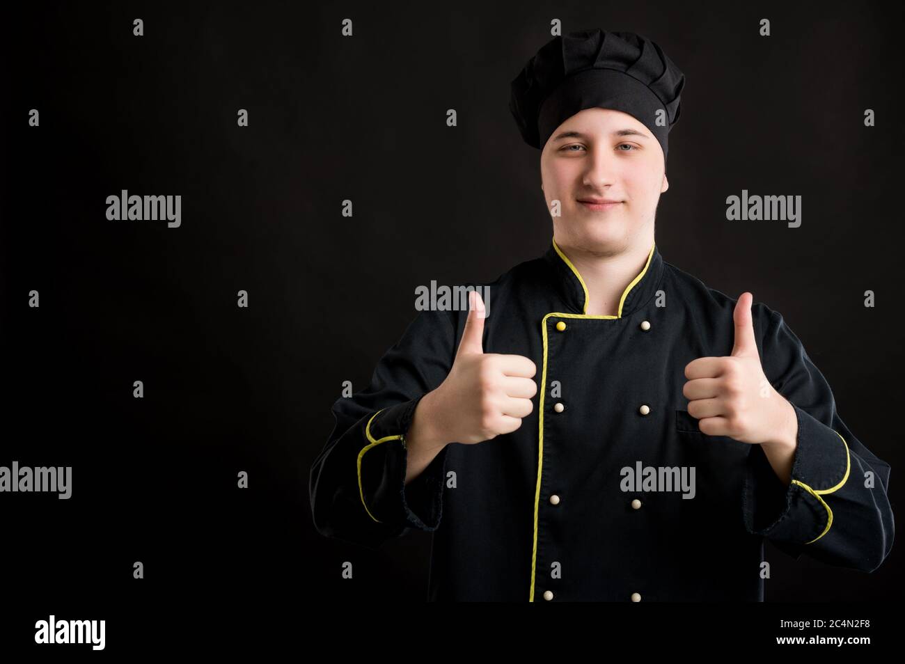 Portrait d'un jeune homme habillé dans un costume de chef noir montrant les pouces vers le haut avec les deux mains posant sur un fond noir isolé avec l'espace de copie annonce Banque D'Images