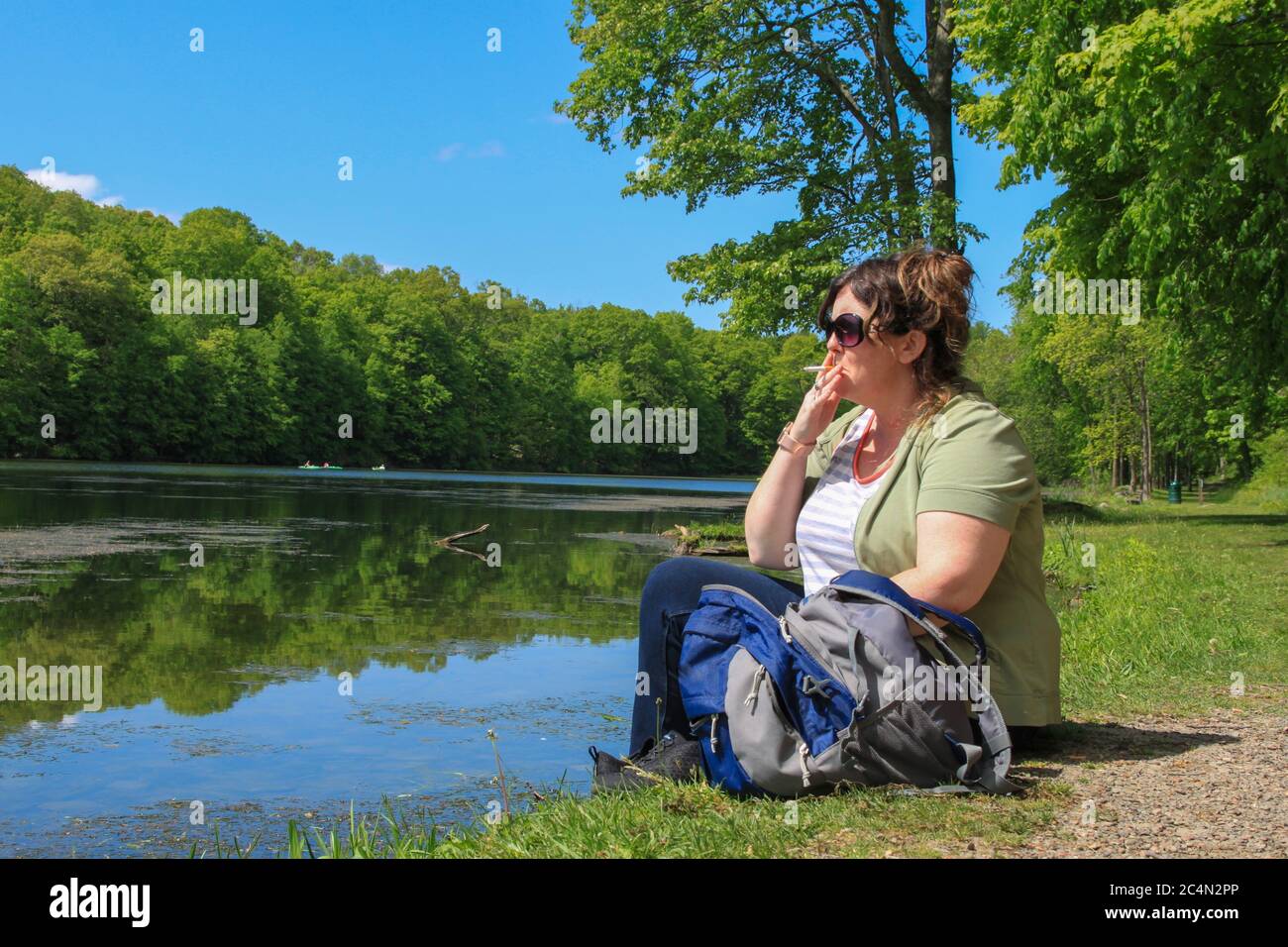 Femme de randonnée assise sur la rive du lac Edge avec sac à dos et Cigarette fumant avec lunettes de soleil Banque D'Images